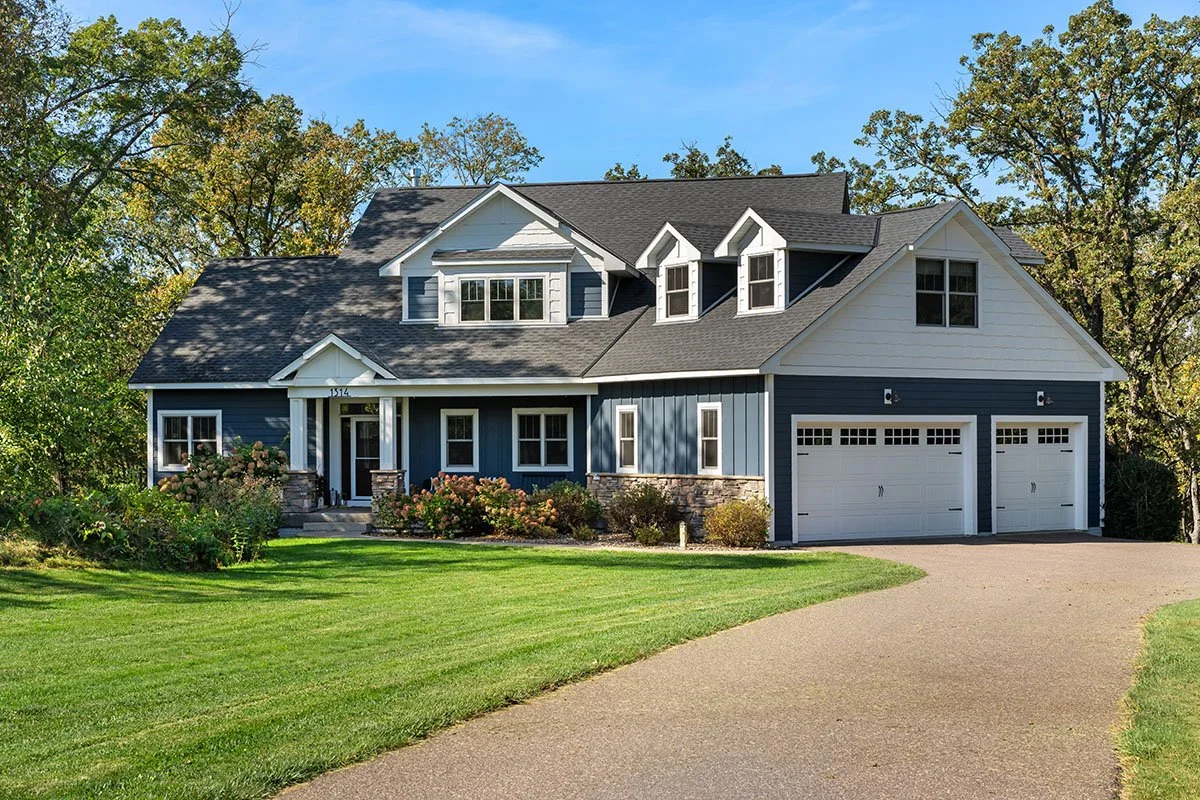 A two-story house with a blue and white exterior, a double garage, and a well-maintained lawn surrounded by trees.