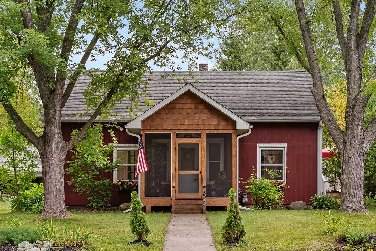 Front view of a red house with a small front porch, two large trees, a concrete walkway, and small shrubs and plants in the yard.