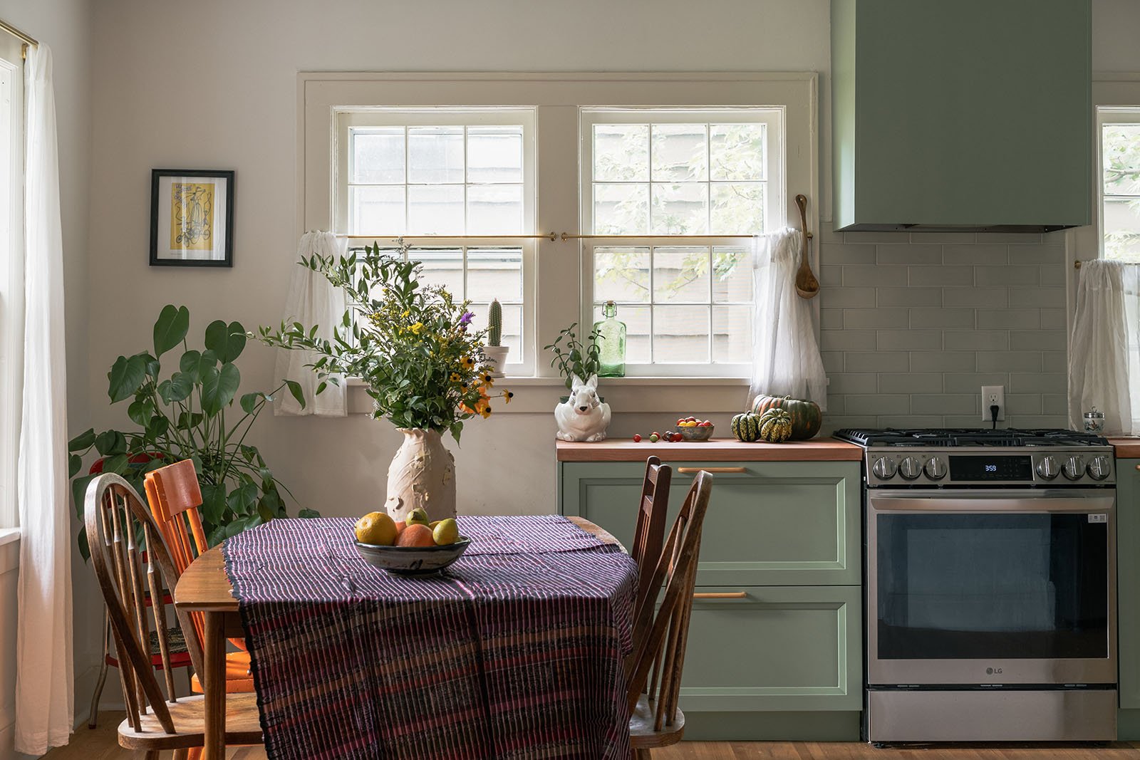 A cozy kitchen with mint green cabinets, a gray stove, and a wooden dining table with four chairs. A large vase with fresh flowers is on the table, and a bowl of fruit is on the tablecloth. There are windows with white curtains and various decorative