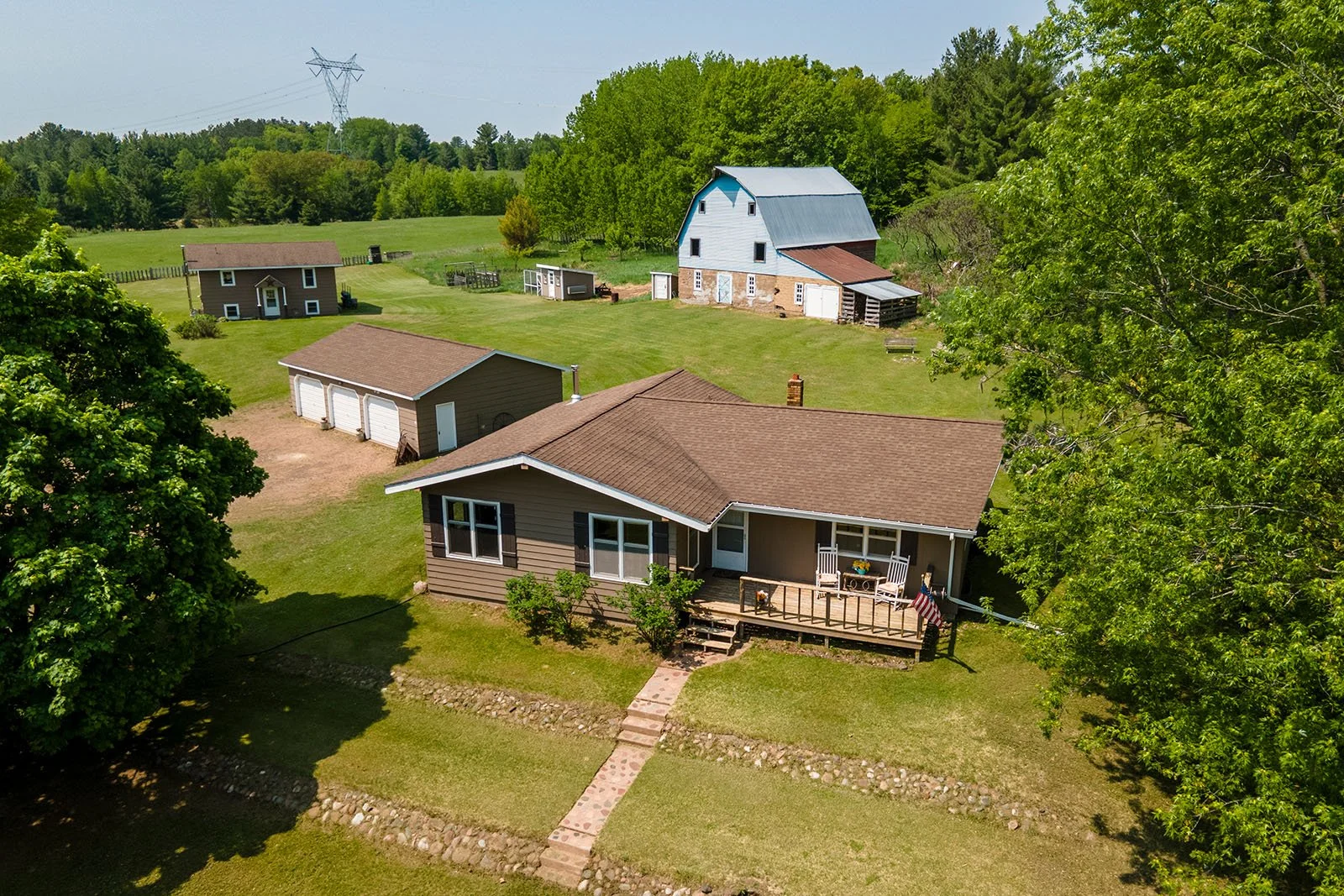 Aerial view of a rural property with a house, a detached garage, a barn, and surrounding green trees and grass fields.