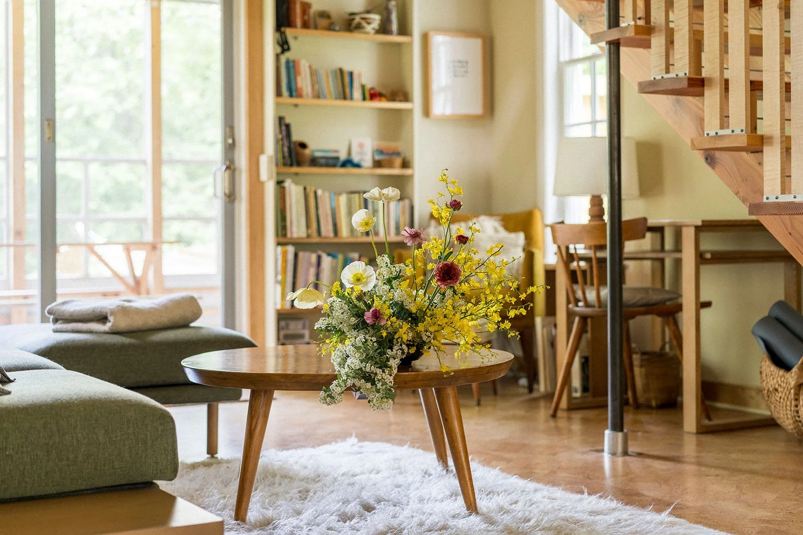 A living room with a glass door, a bookshelf filled with books, a yellow armchair, a small wooden dining table with a wicker chair, a staircase with wooden steps, and a round wooden coffee table with a bouquet of white, pink, yellow, and purple flowe