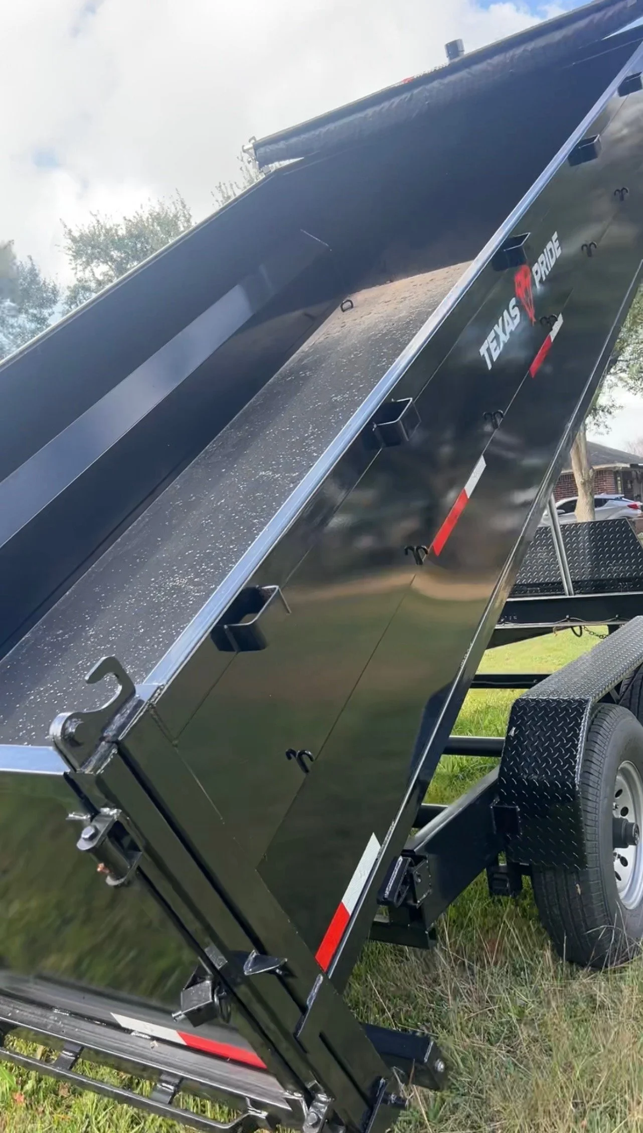 A black Texas Pride utility trailer with a tilt-up bed, positioned on grass in an outdoor setting, with trees and a partly cloudy sky in the background.