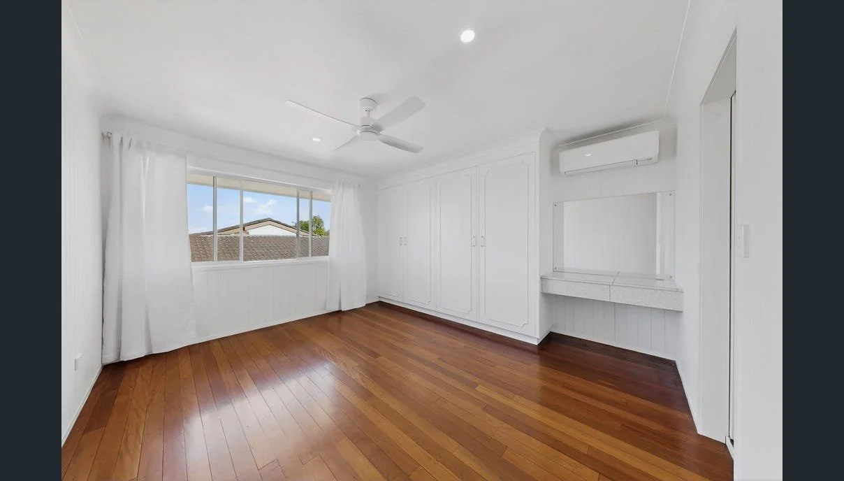 Empty bedroom with hardwood floors, white walls, a large window with curtains, an air conditioning unit, a ceiling fan, and built-in white wardrobe.