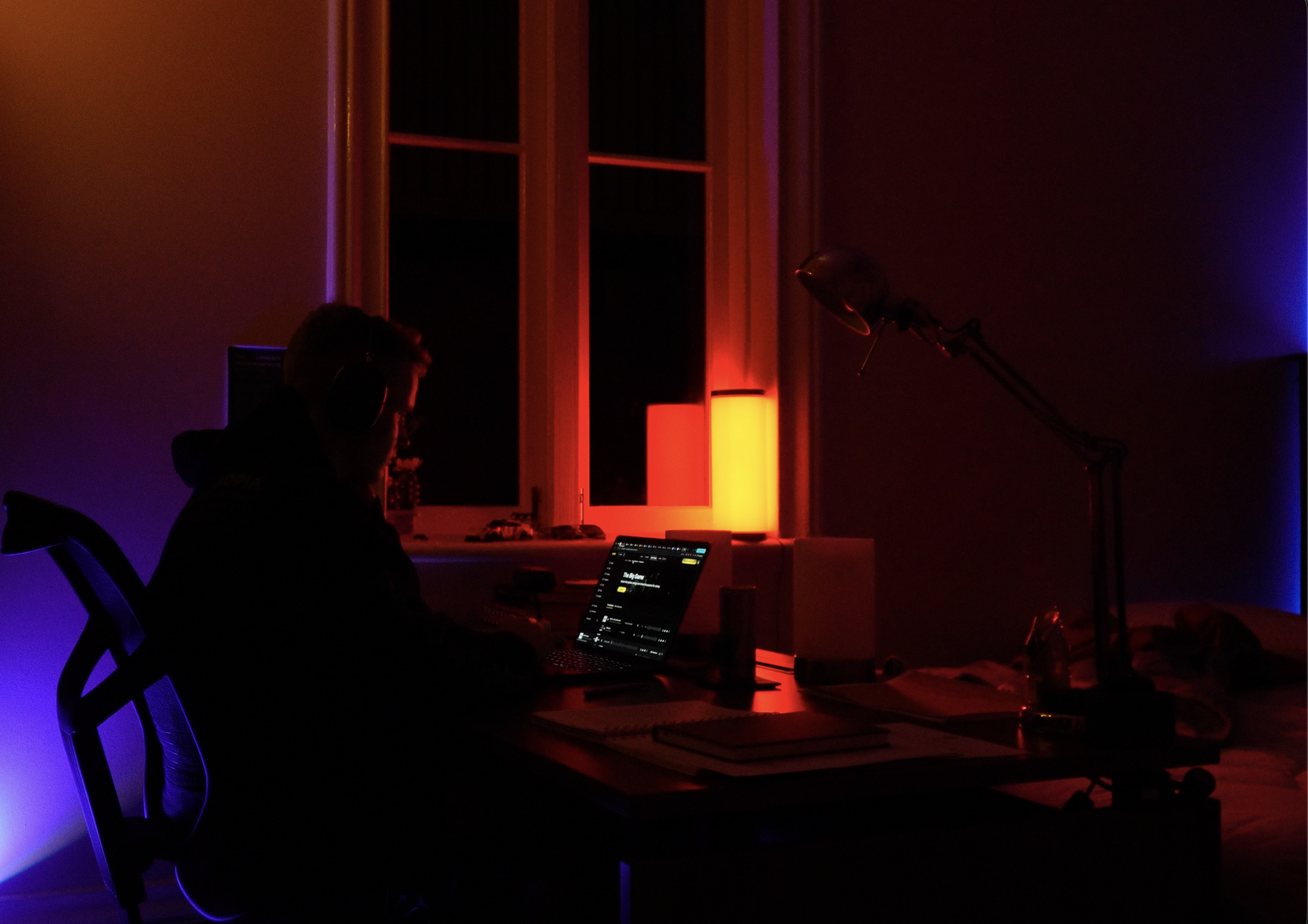 A person sitting at a desk in a dimly lit room illuminated by colorful LED lights, working on a laptop with headphones on.