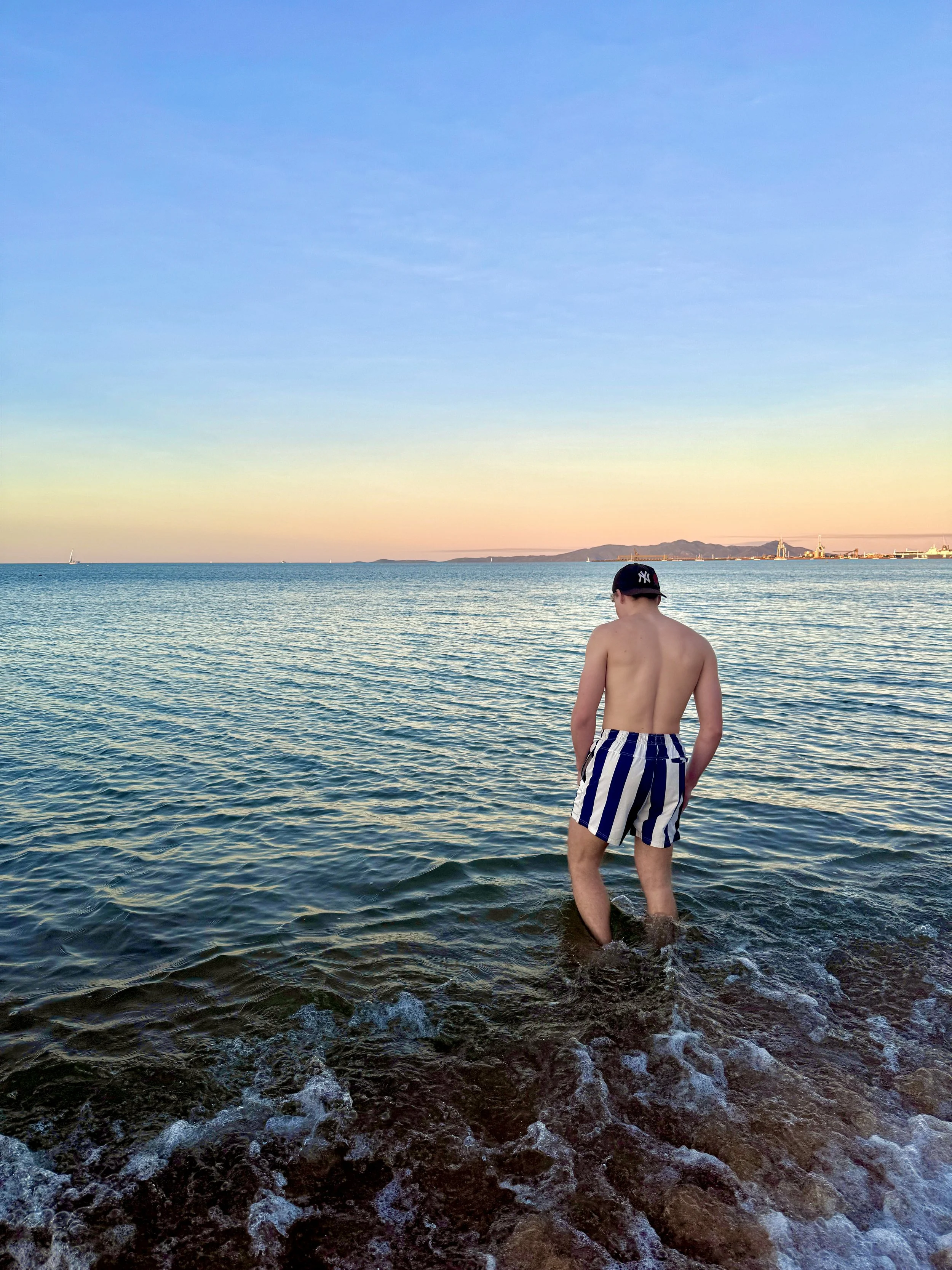 A person standing knee-deep in water at the beach during sunset, wearing striped shorts and a cap.