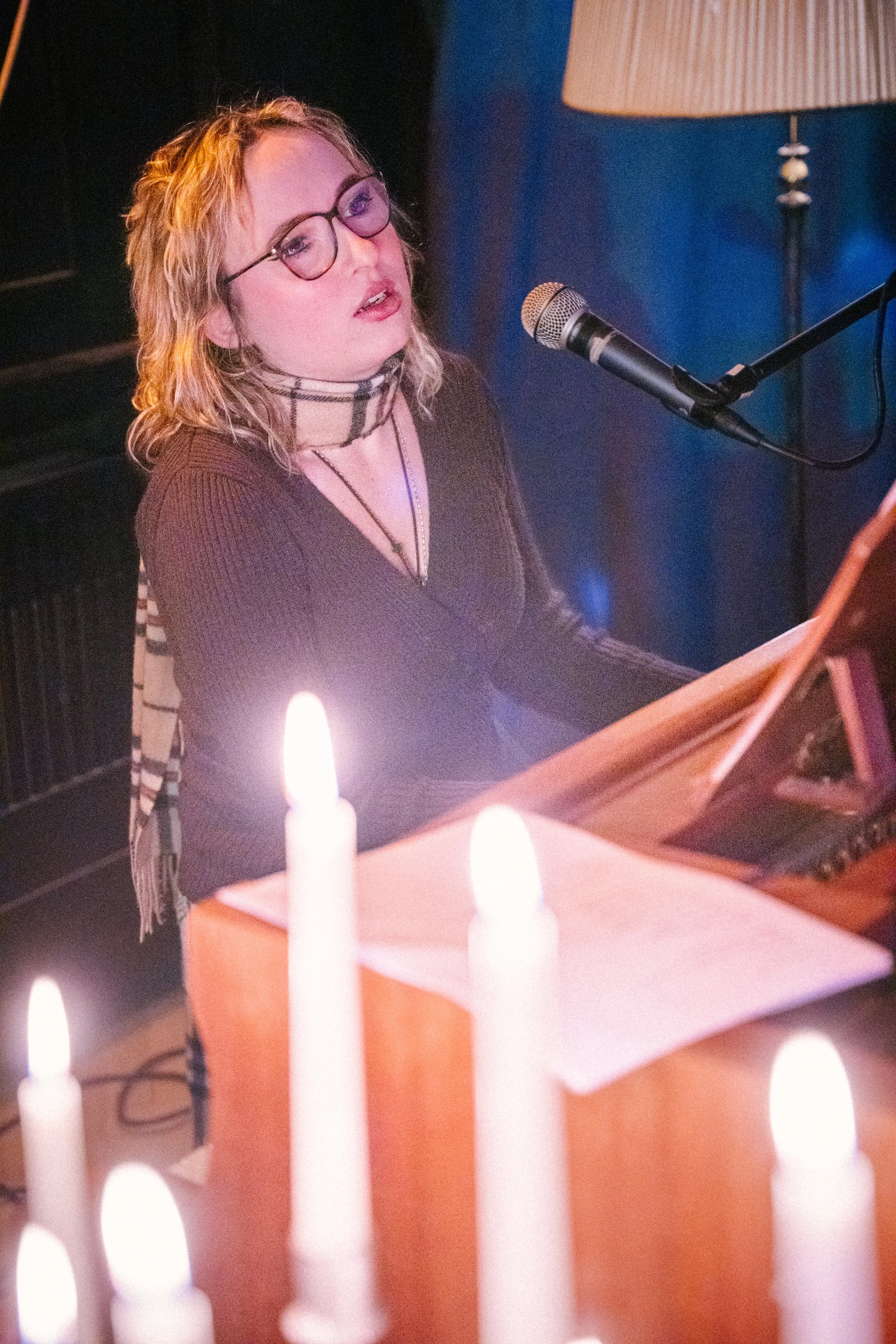 Eine Frau mit Brille singt oder spielt auf einem Klavier, umgeben von Kerzenlicht, in einem gemütlichen Raum.