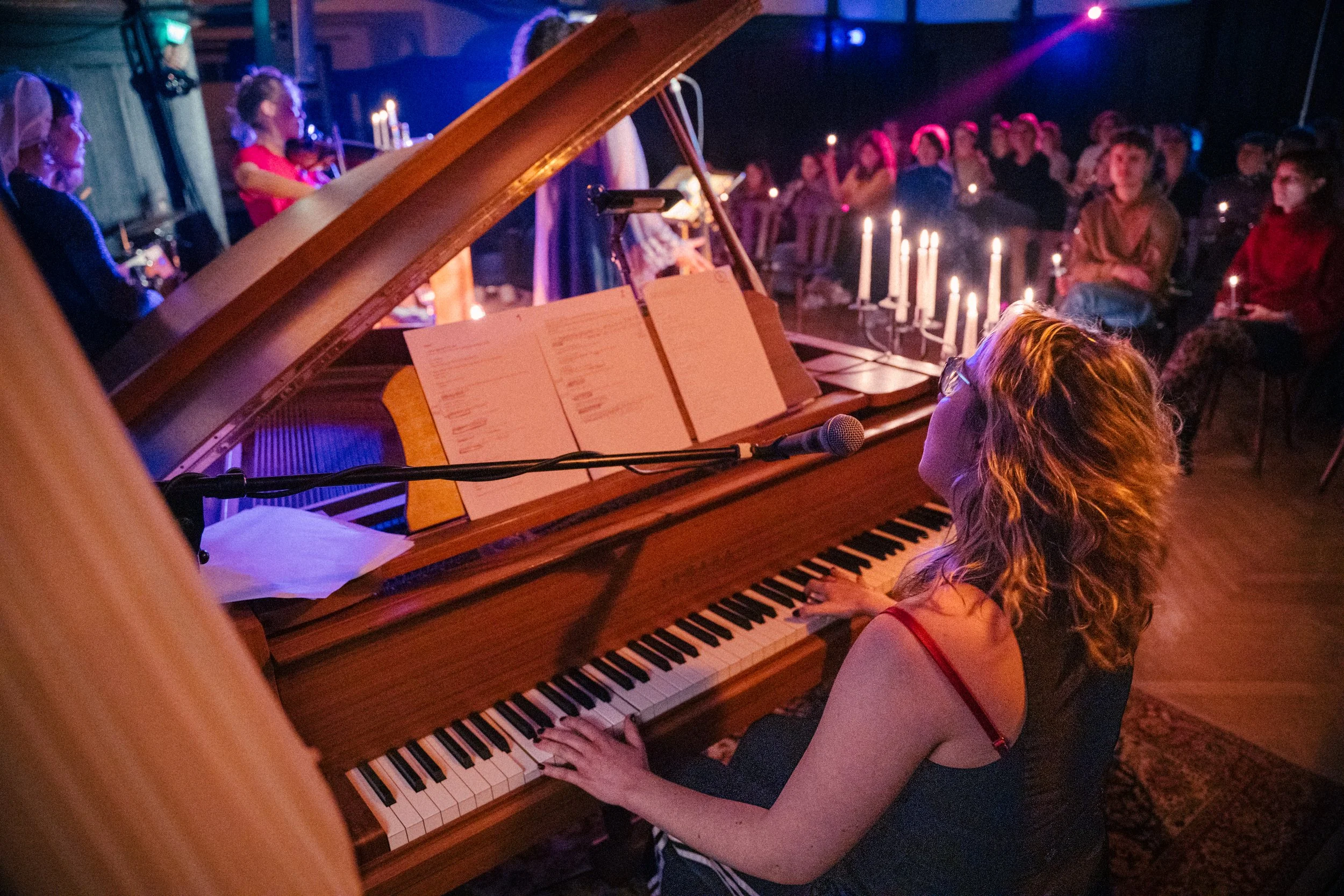Eine Frau spielt Klavier bei einem Live-Konzert, im Hintergrund sitzen das Publikum und Kerzenlicht.