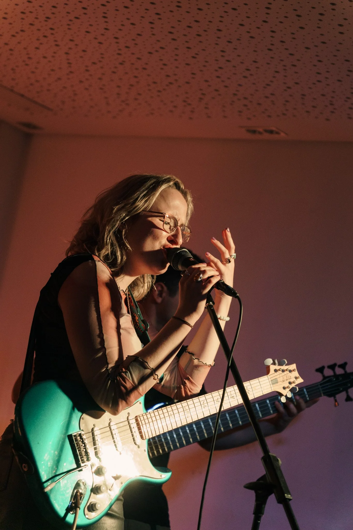 Eine Frau mit Brille singt in einem Mikrofon und spielt eine grüne E-Gitarre, während sie auf einer Bühne auftritt.