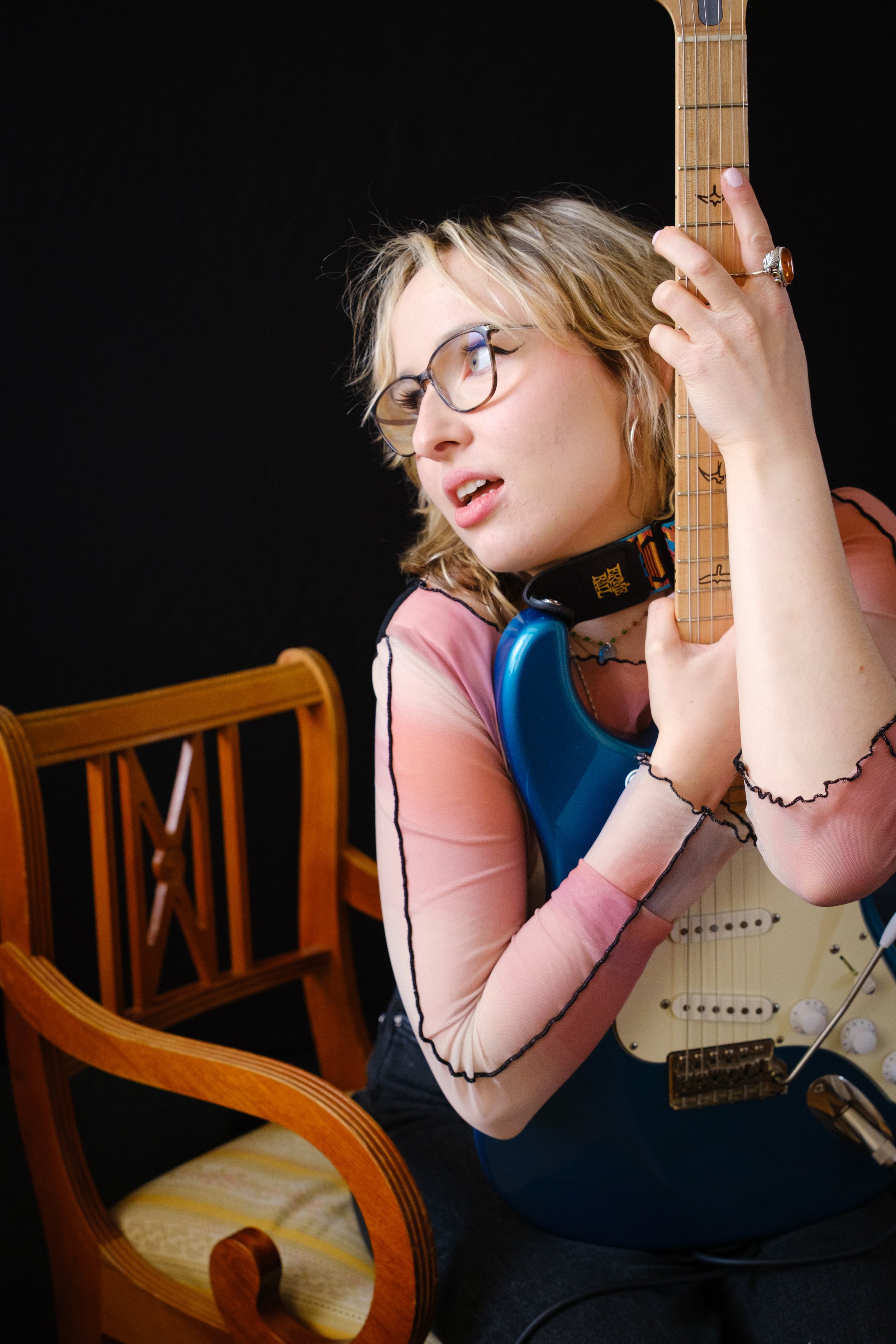 Junge Frau mit Brille und blondem Haar spielt E-Gitarre, sitzt auf Stuhl, vor dunklem Hintergrund.