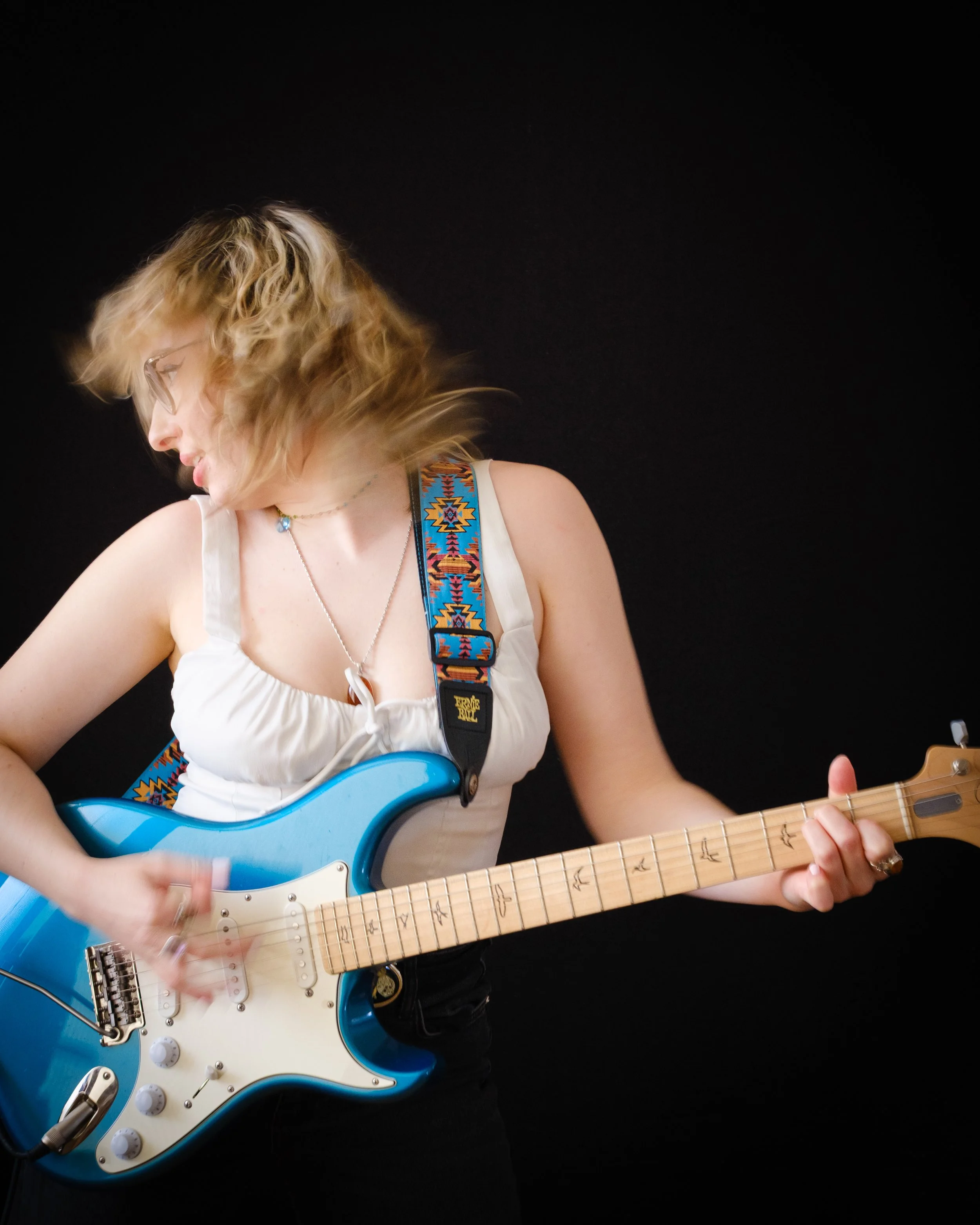 Junge Frau spielt E-Gitarre vor schwarzem Hintergrund, trägt weiße Top und farbenfrohen Gitarrengurt, hat kurze gelockte Haare und trägt Brille.