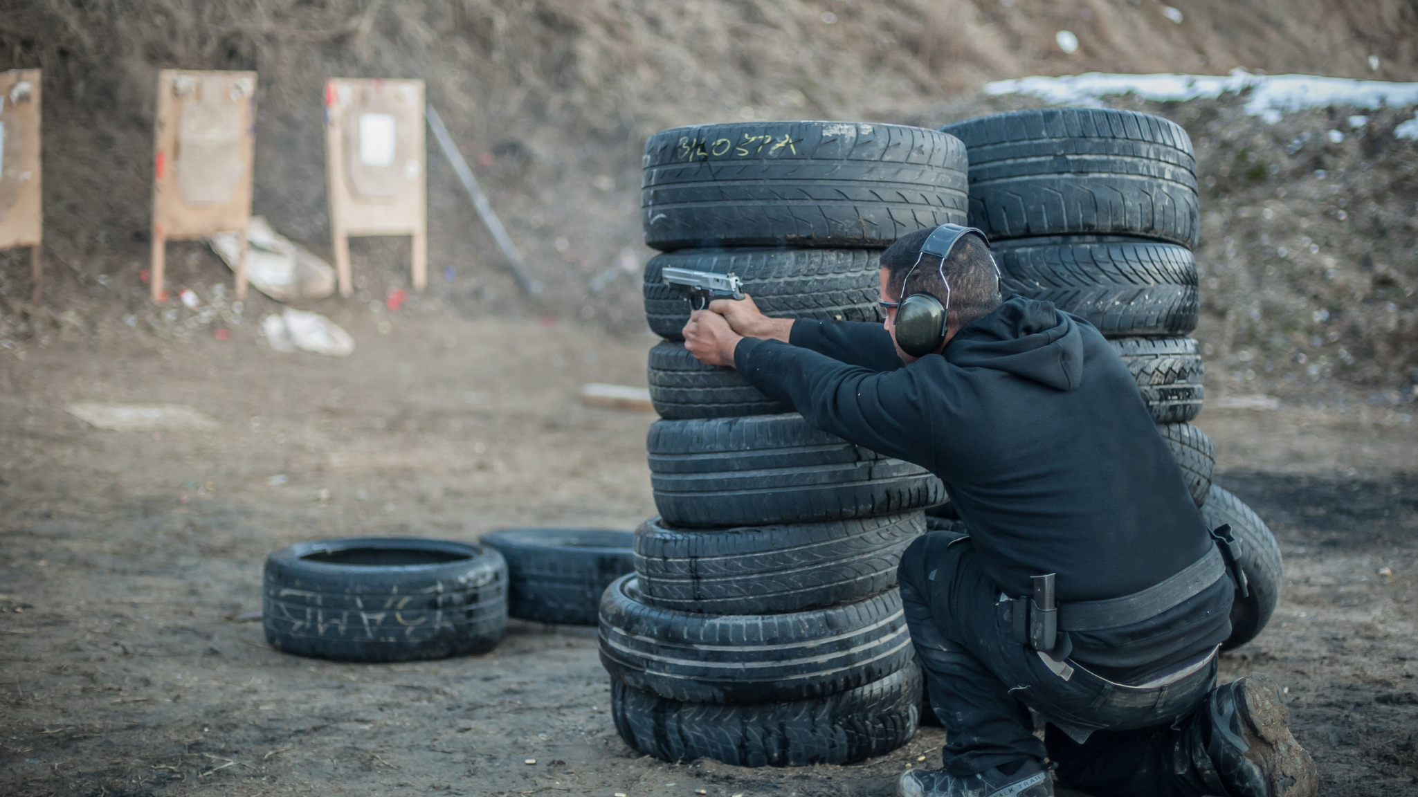 A man kneeling behind a stack of tires at a shooting range, aiming a handgun, wearing hearing protection and sunglasses.