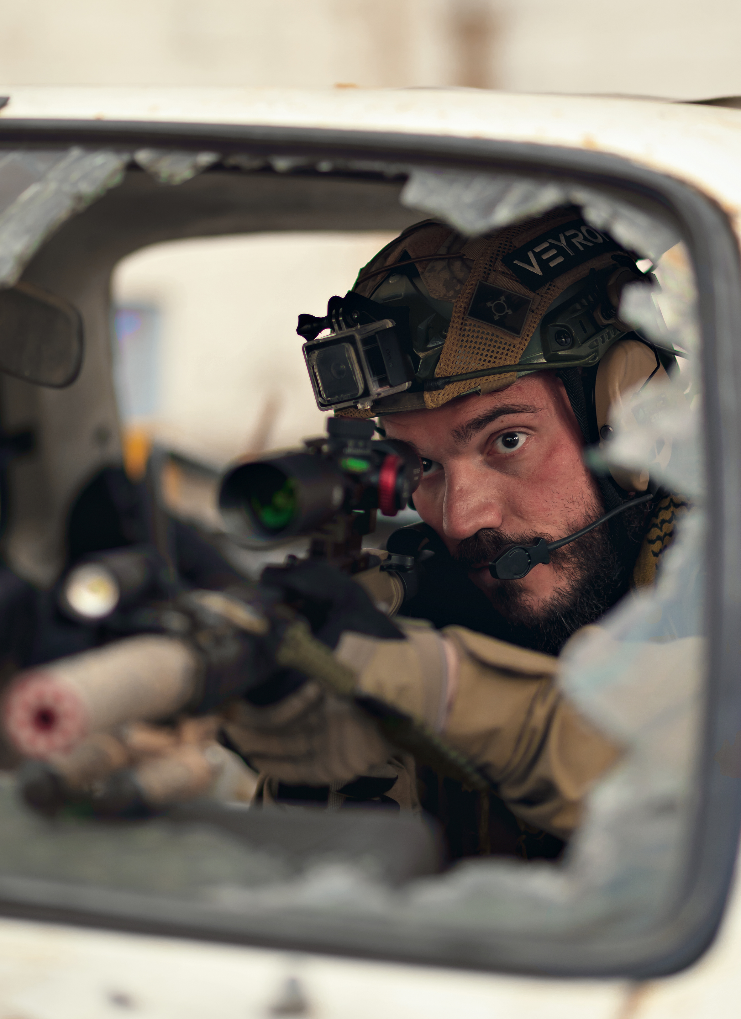 A soldier aiming a sniper rifle through a broken window in a building, wearing desert camouflage and tactical gear.
