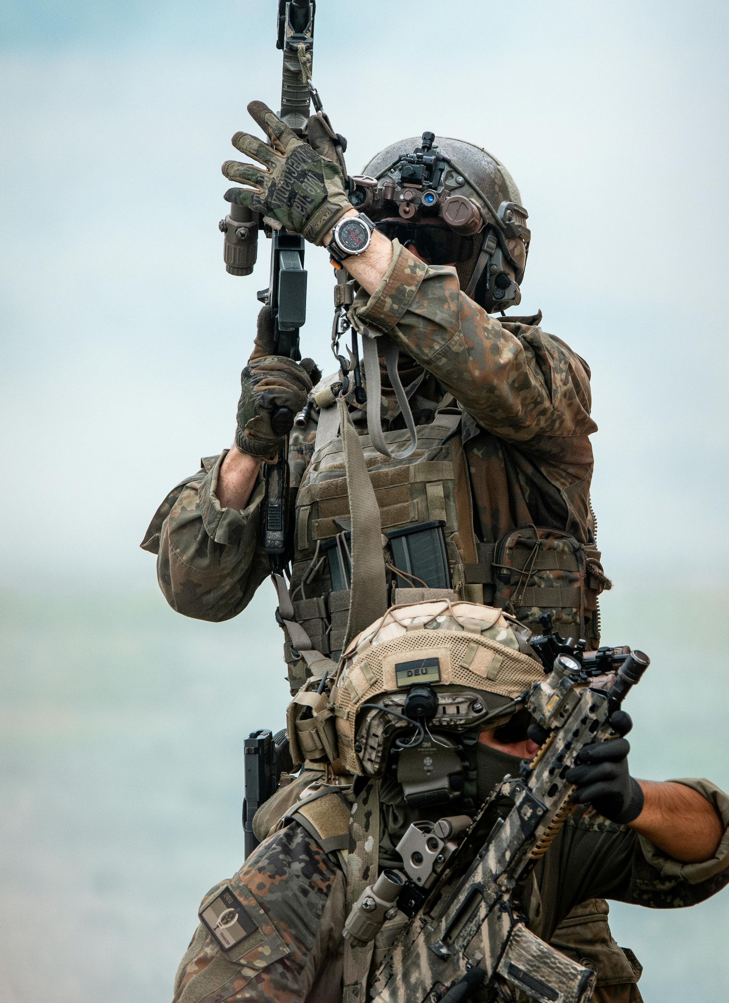 Two soldiers in camouflage gear with helmets and tactical equipment, holding rifles, one standing and one kneeling, outdoors in a cloudy sky.
