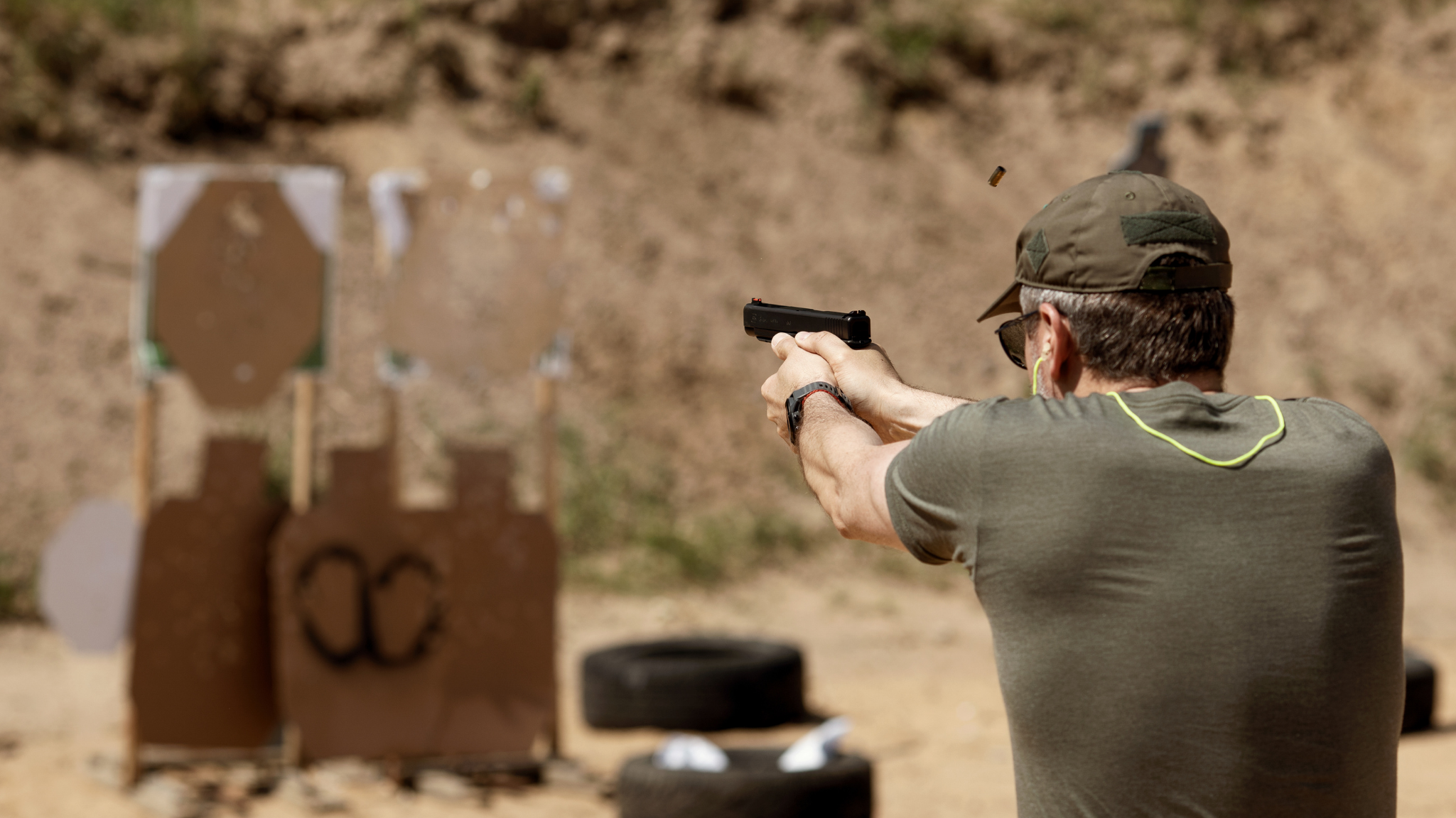 Man at outdoor shooting range aiming a handgun at targets, with the targets and tires in the background.