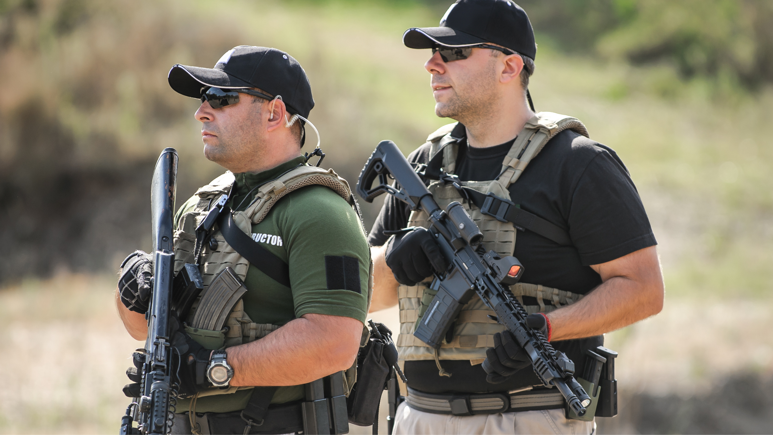 Two armed men wearing tactical gear, sunglasses, and hats standing outdoors in a grassy area.
