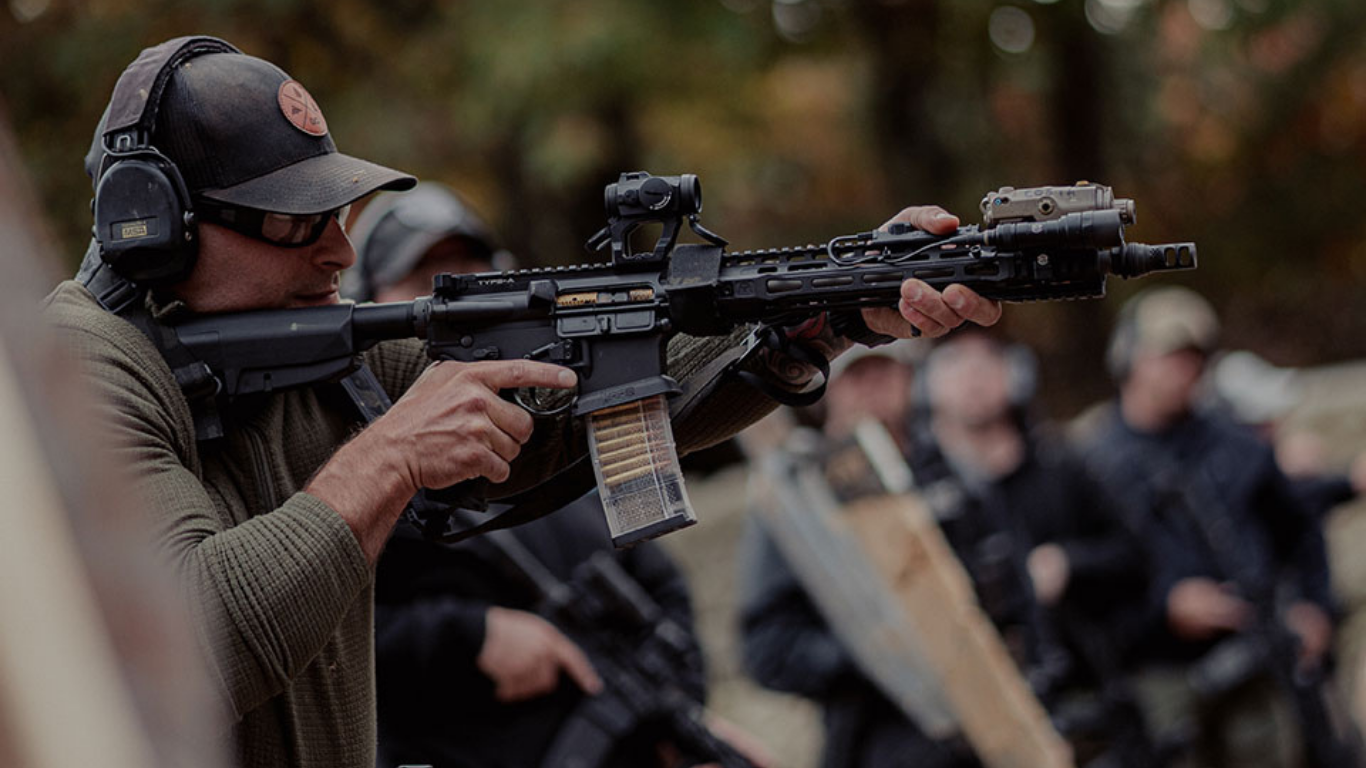A person wearing a cap, safety glasses, and hearing protection aiming a rifle during a shooting practice or training session.