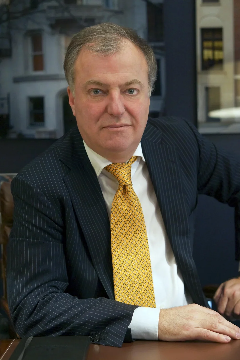 A middle-aged man with gray hair and blue eyes, wearing a dark pinstripe suit, white shirt, and yellow textured tie, sitting at a desk with buildings visible in the background.