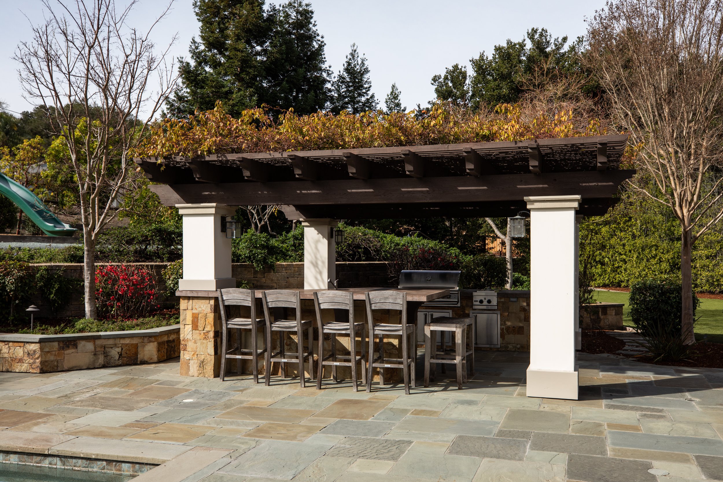 Outdoor patio with a covered bar area, stone counter, wooden bar stools, and a built-in grill, surrounded by landscaped yard with trees and bushes.