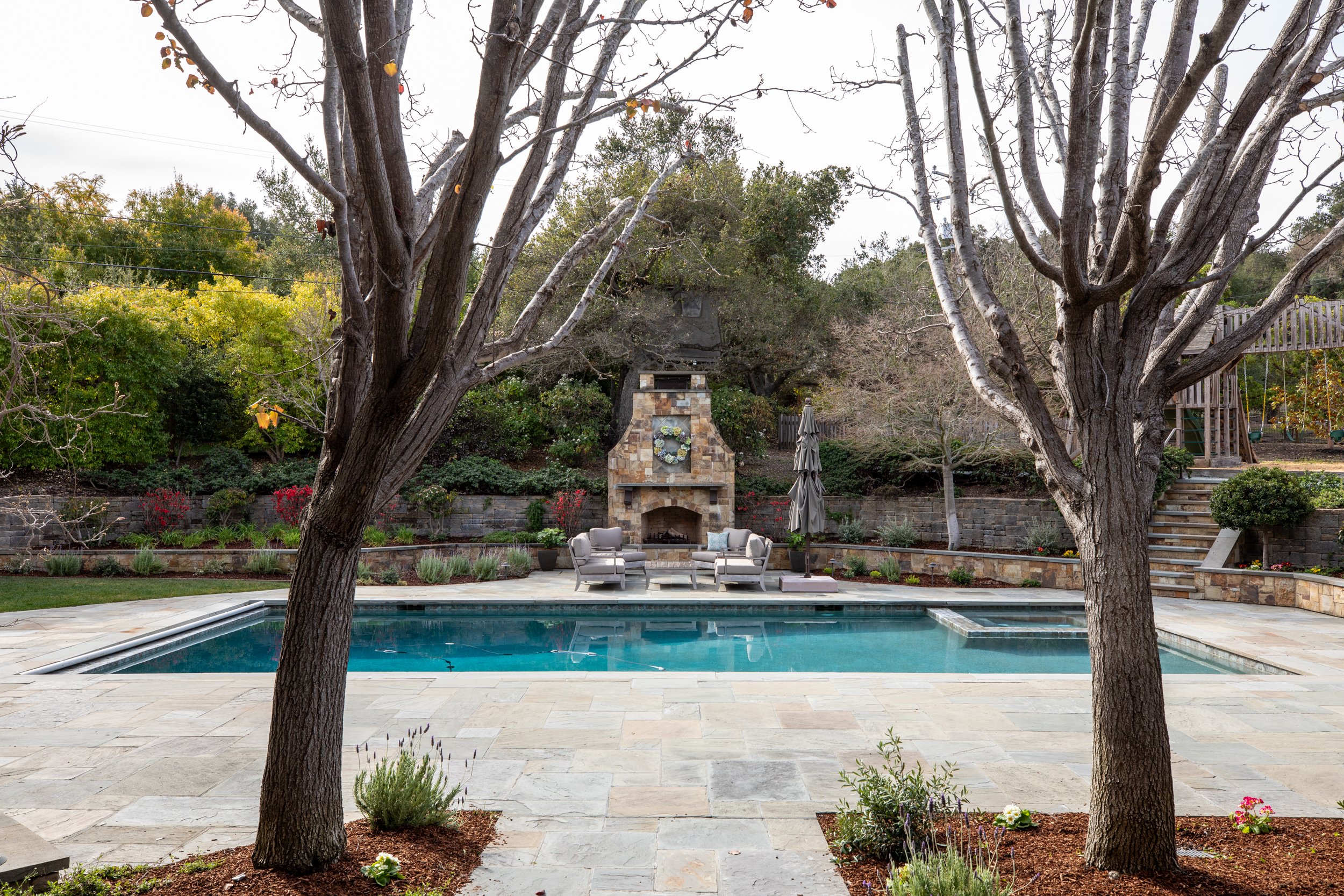 Backyard with a swimming pool, fireplace, and outdoor seating surrounded by trees and landscaped garden.