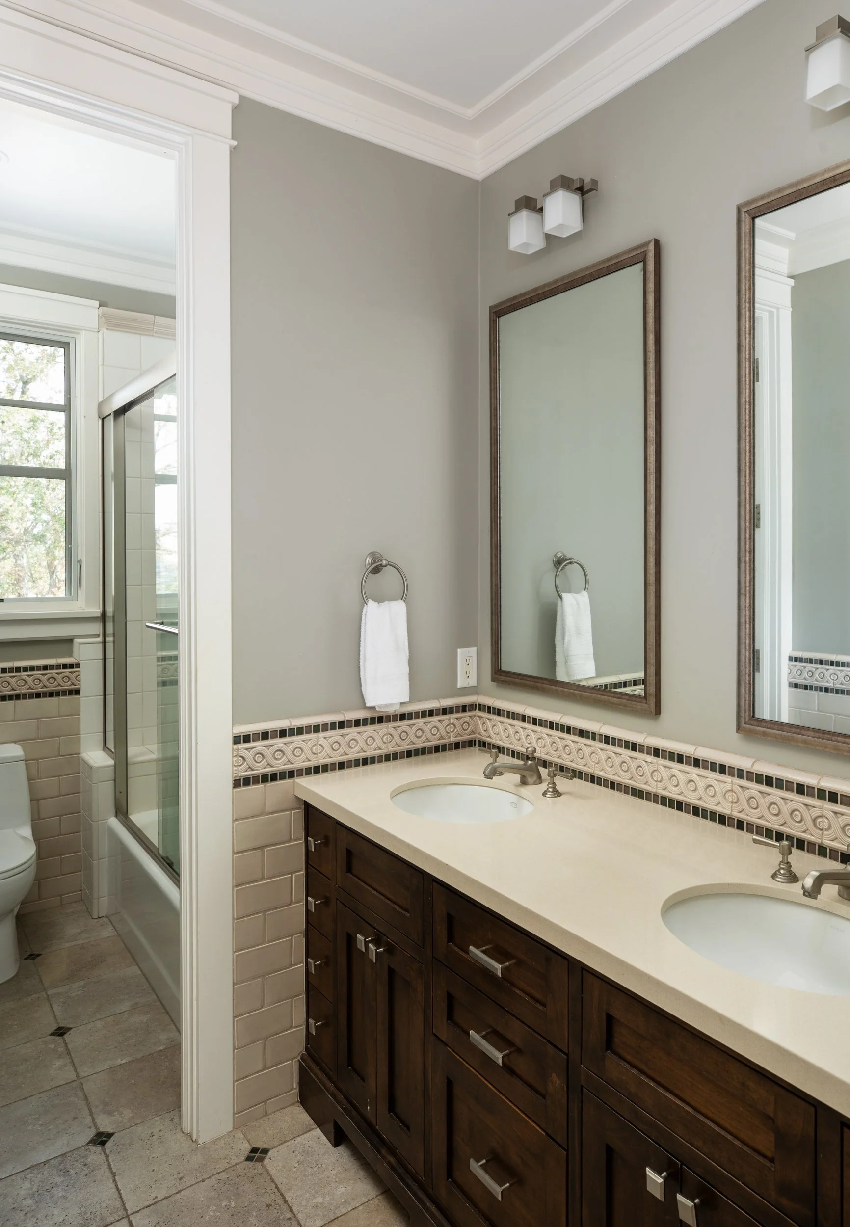 Bathroom with double sink vanity, large mirror, gray walls, towel rings with white towels, wall-mounted light fixtures, and partially visible shower and toilet area.