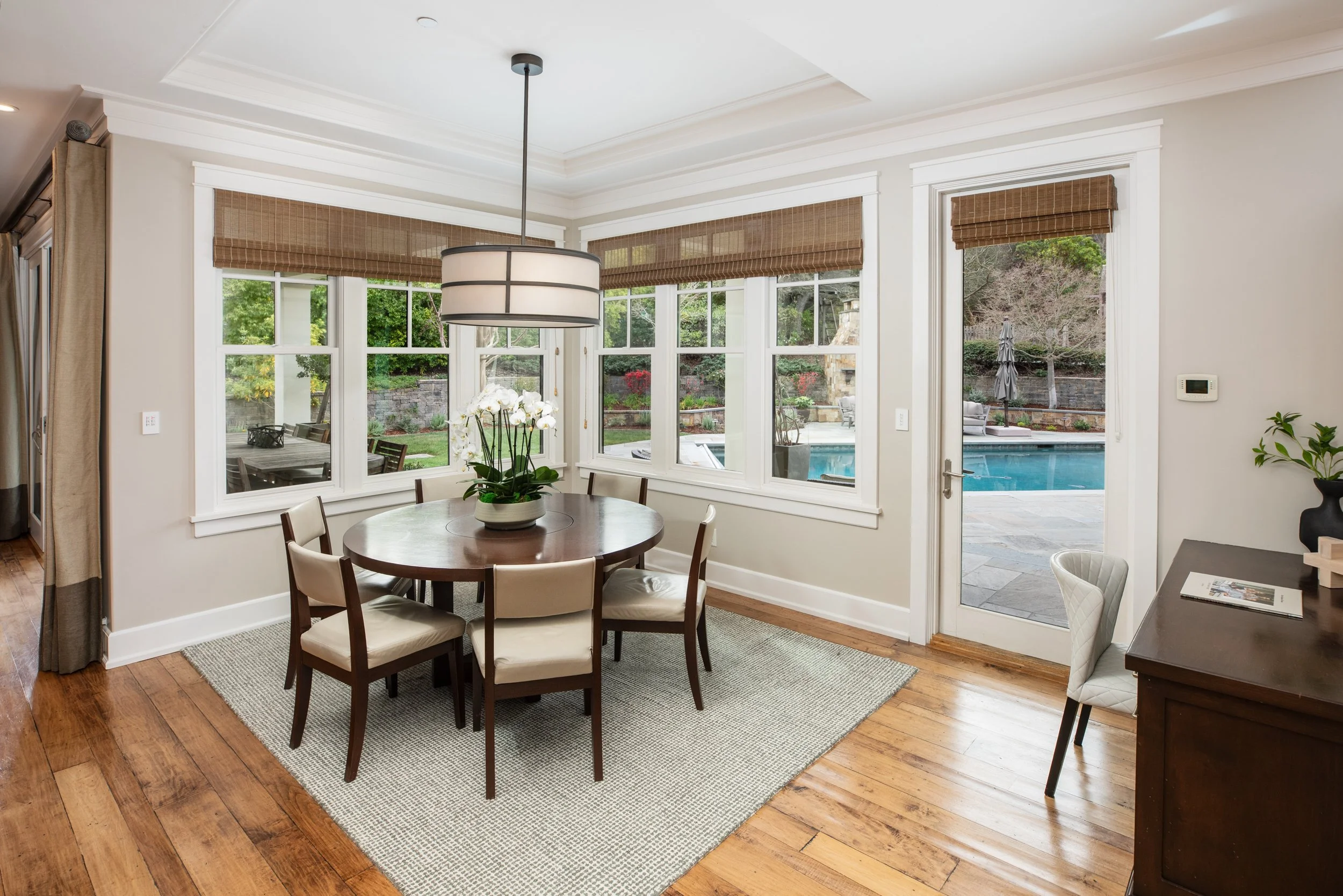 Dining area with a round wooden table, six chairs, a potted orchid, and a large pendant light, with multiple windows showing an outdoor patio and pool.