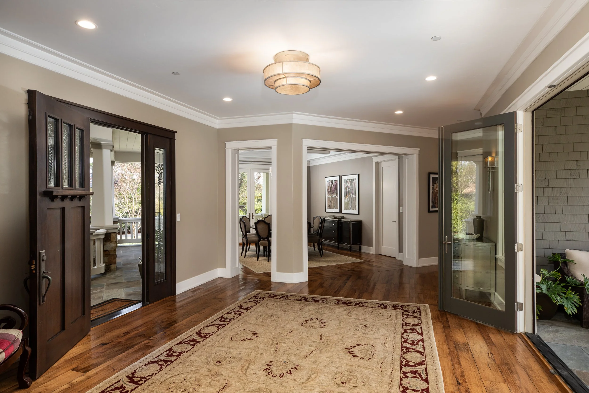 Living room with hardwood floors, a large area rug, and doorways leading into dining and outdoor spaces. Ceiling light fixture and recessed lighting are visible.