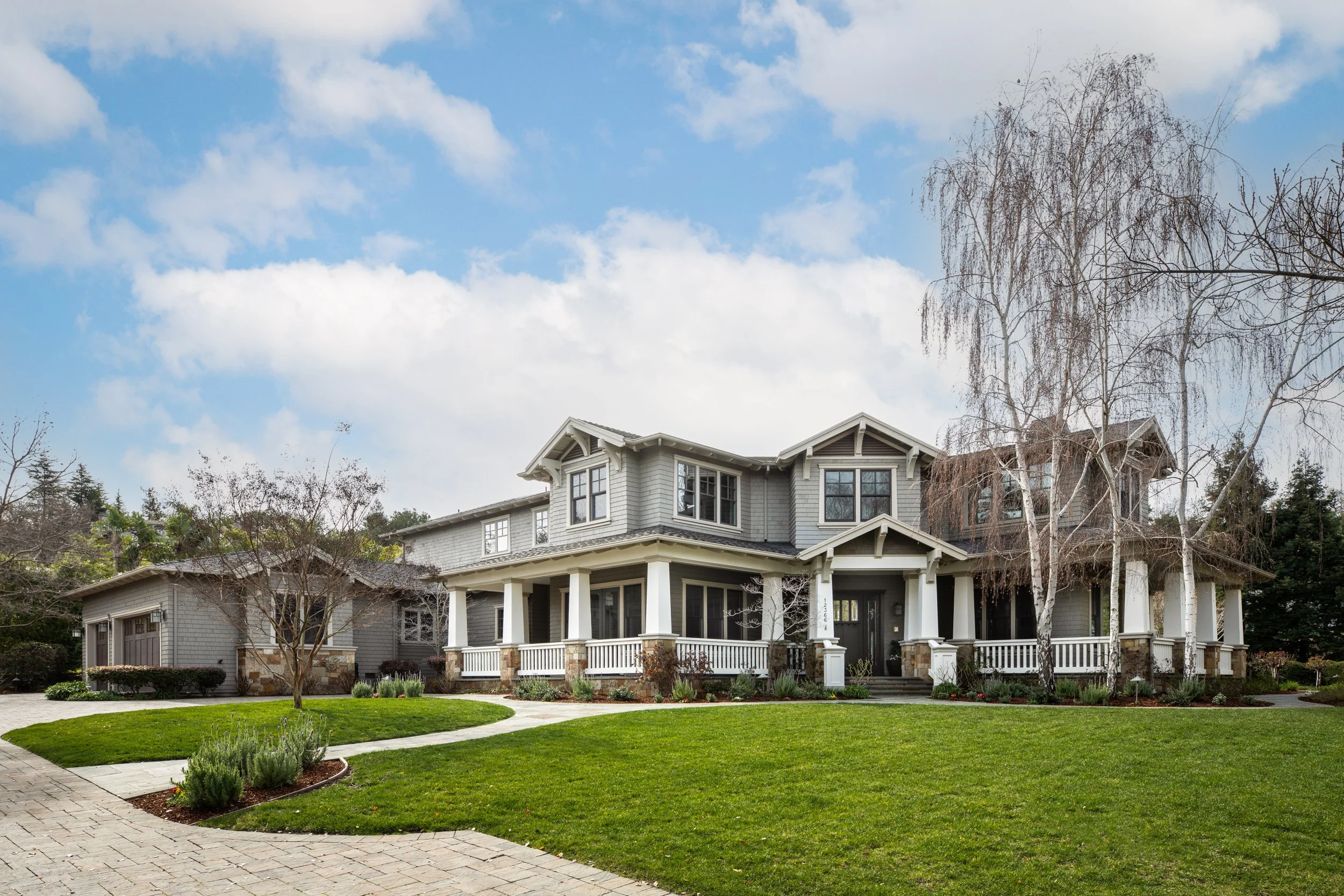 A large, two-story house with gray siding, white columns, and a front porch. The property has a well-maintained lawn with curved pathways and a few leafless trees, under a partly cloudy sky.