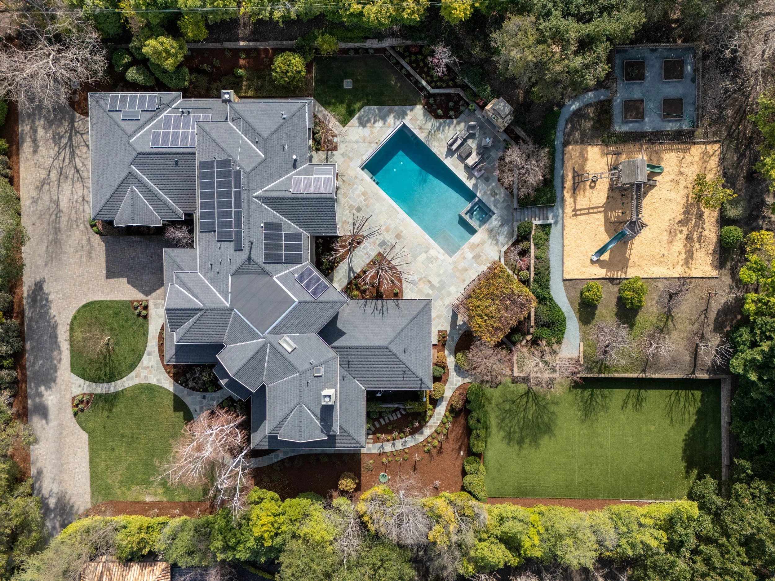 Aerial view of a house with a swimming pool, a playground area, and a well-maintained lawn surrounded by trees.