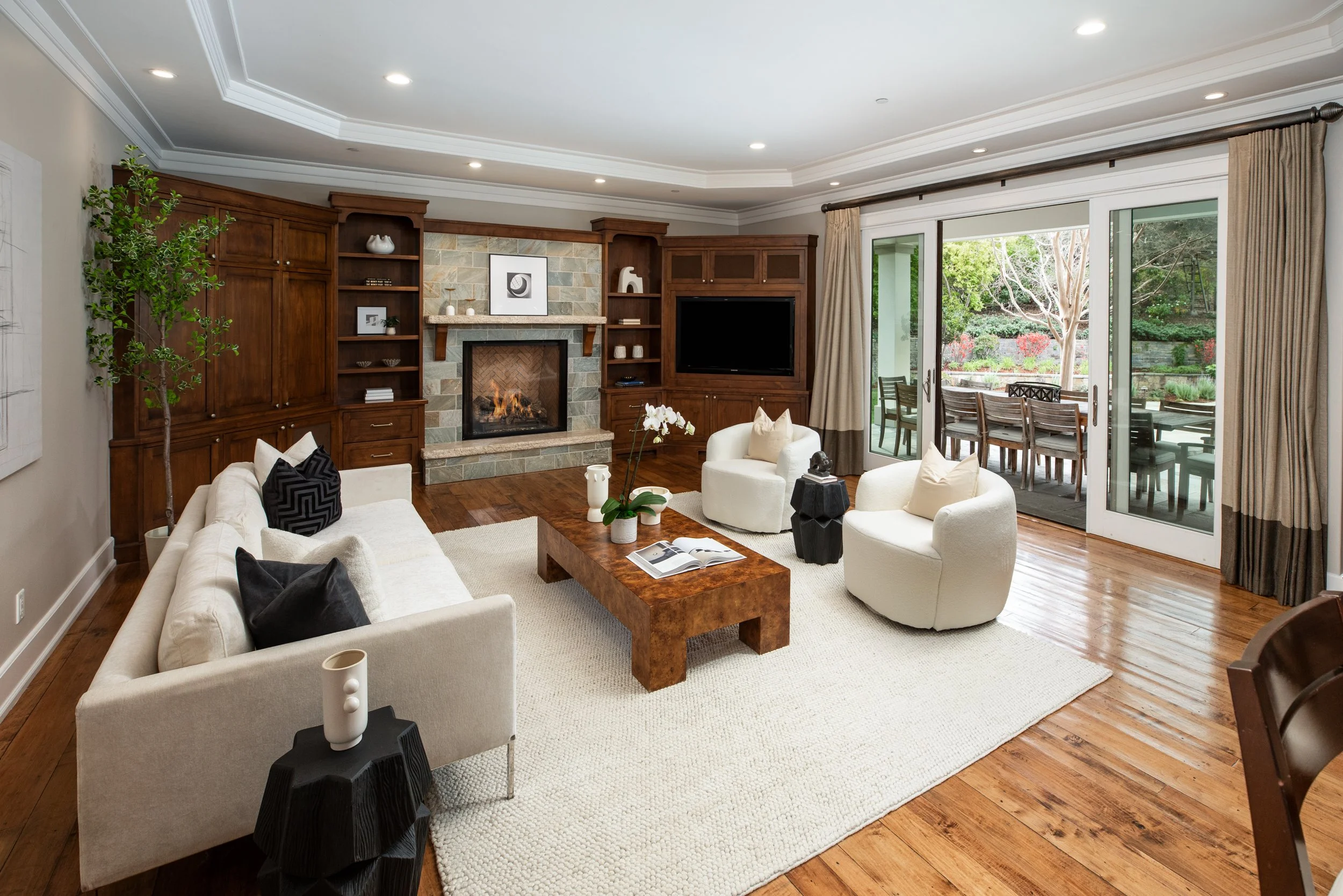 Living room with white sofas, two white armchairs, a wooden coffee table with a book on top, a fireplace with stone surround, a built-in wooden entertainment center with a black TV, large sliding glass doors leading to outdoor patio with dining table