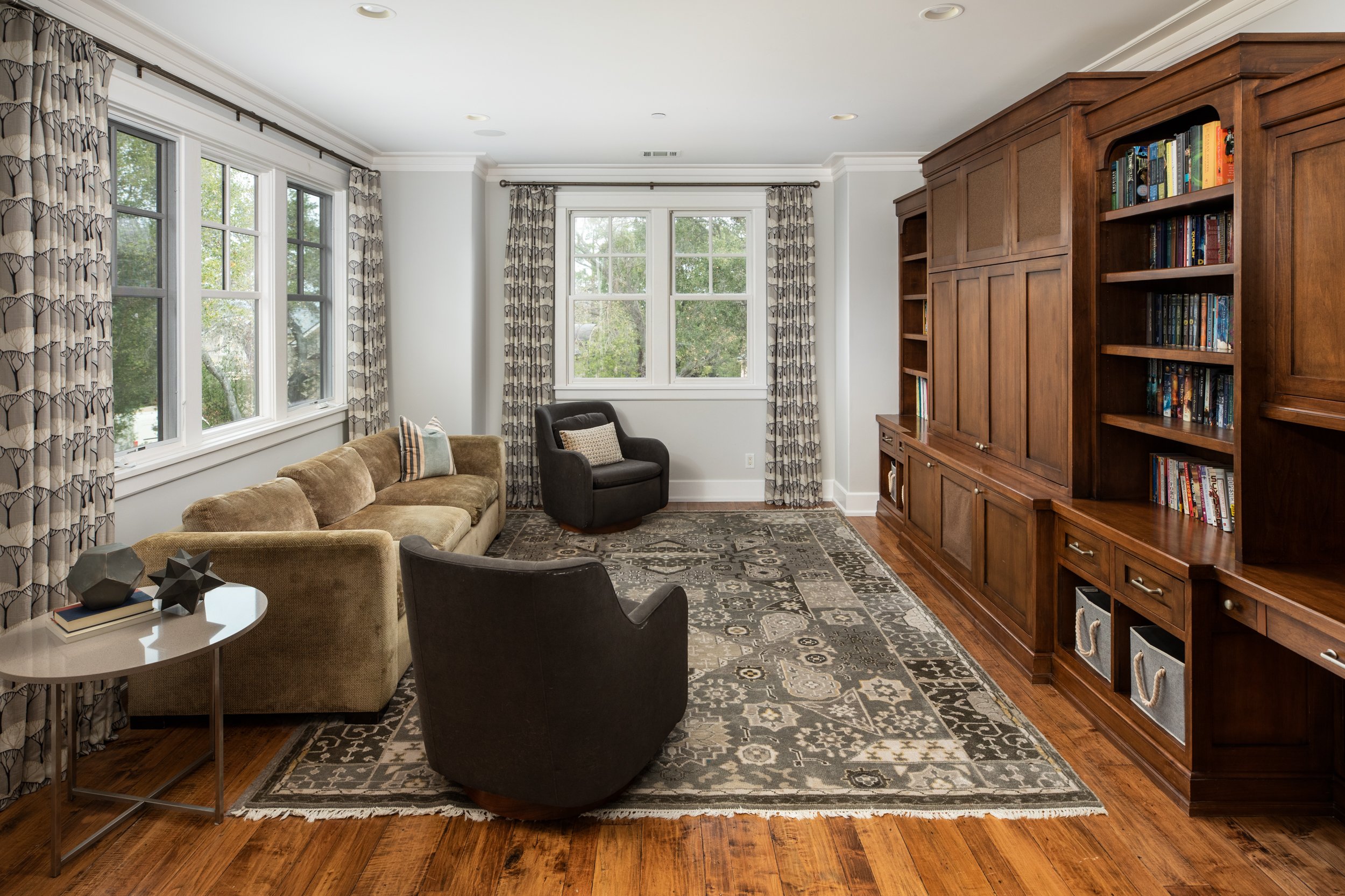 Living room with large windows, patterned curtains, beige sofa, black armchair, area rug, and wooden wall unit with shelves and drawers.