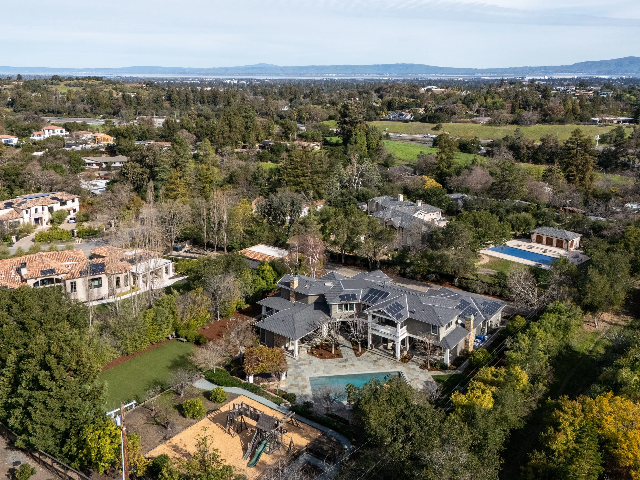 Aerial view of a residential neighborhood with houses, trees, and a swimming pool, with a distant landscape and mountains in the background.