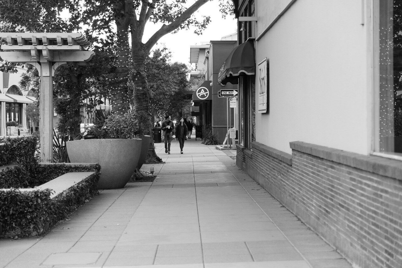 Black and white photo of a sidewalk in an urban shopping area with two people walking and talking, storefronts with awnings and signs, large trees with string lights, potted plants, and a building with brick and white walls.