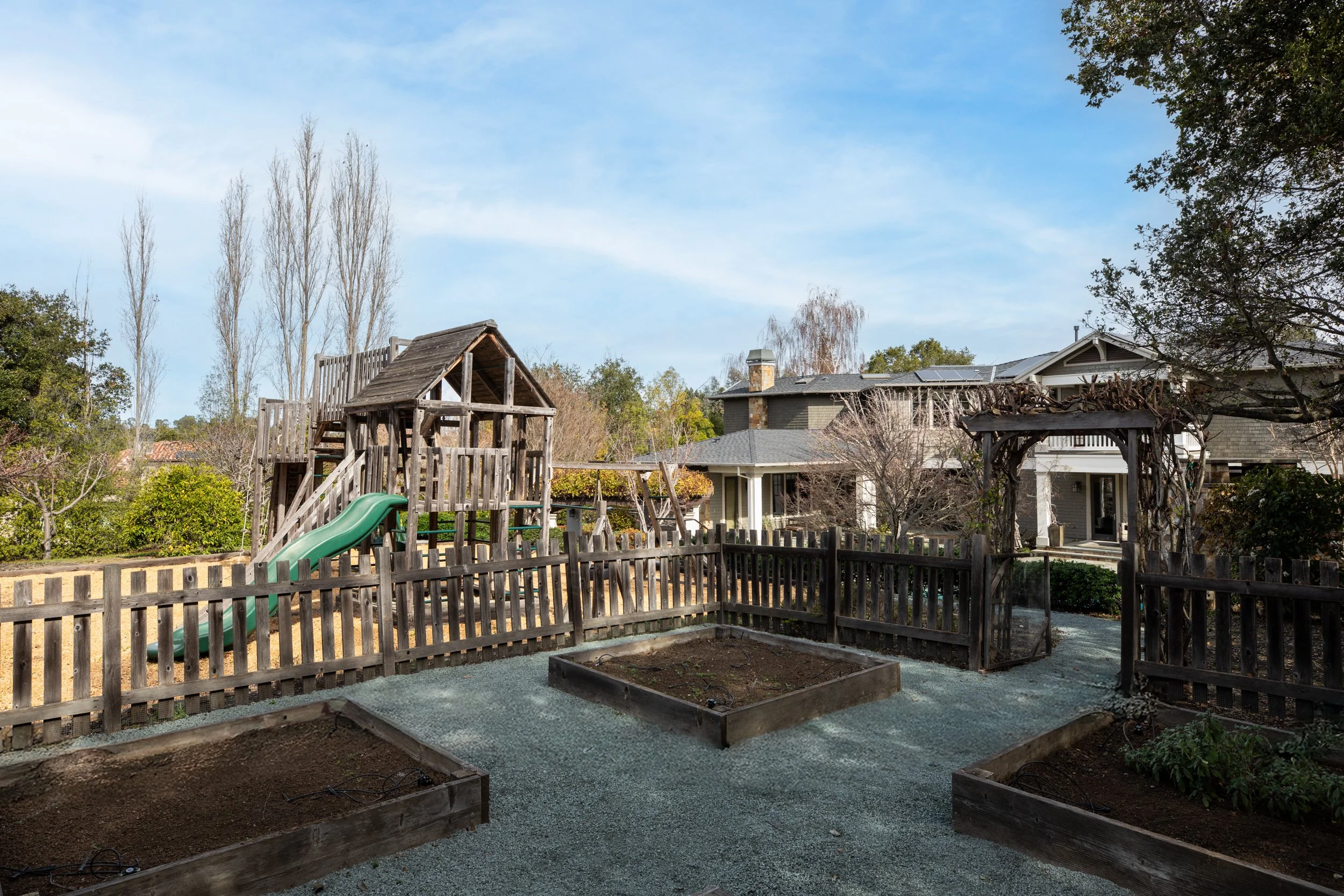 A backyard with a wooden playground structure, a green slide, and a garden bed, surrounded by a wooden fence and neighboring houses during the daytime.