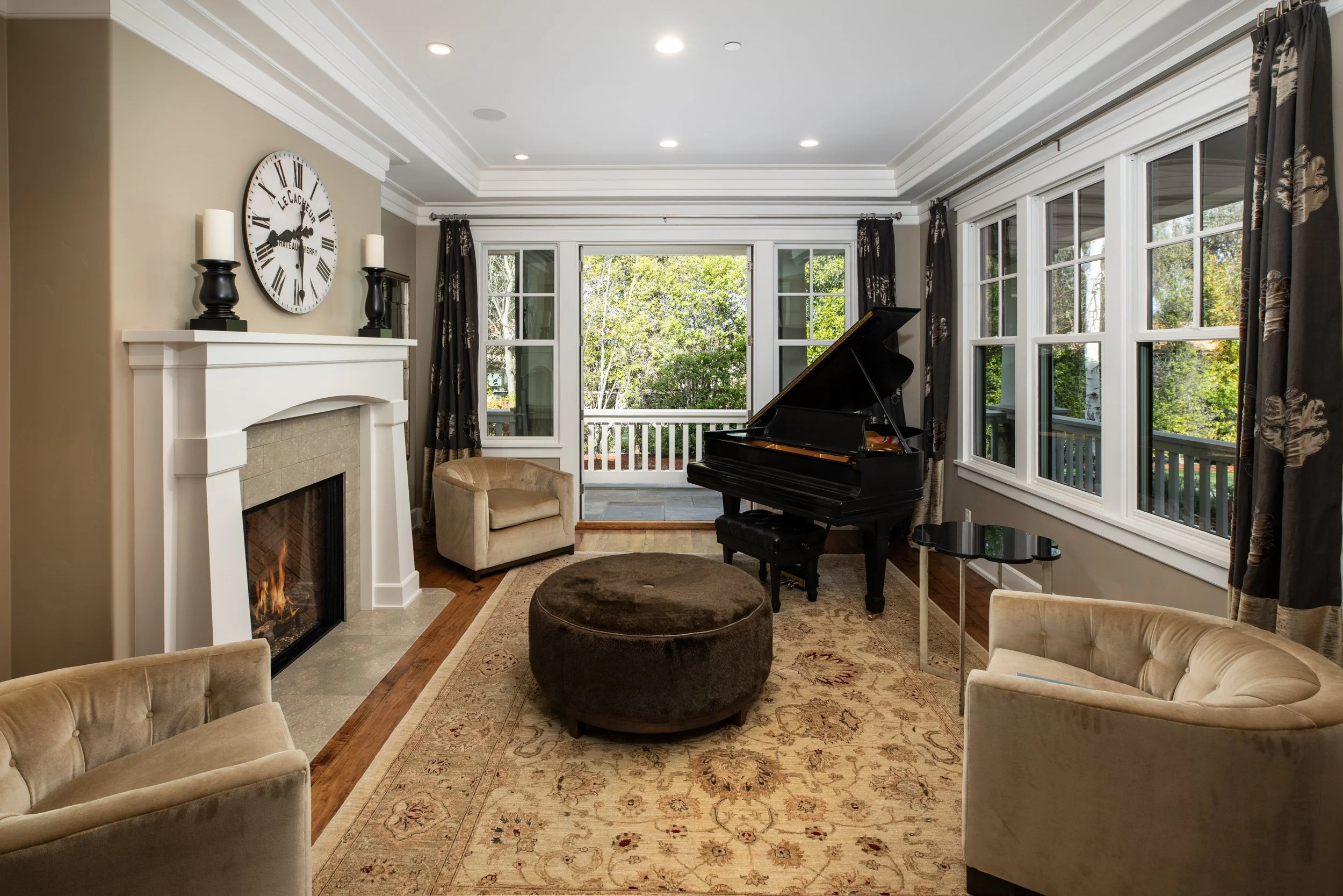 Living room with a fireplace, beige armchairs, a black grand piano, a patterned area rug, and large windows showing outdoor greenery.