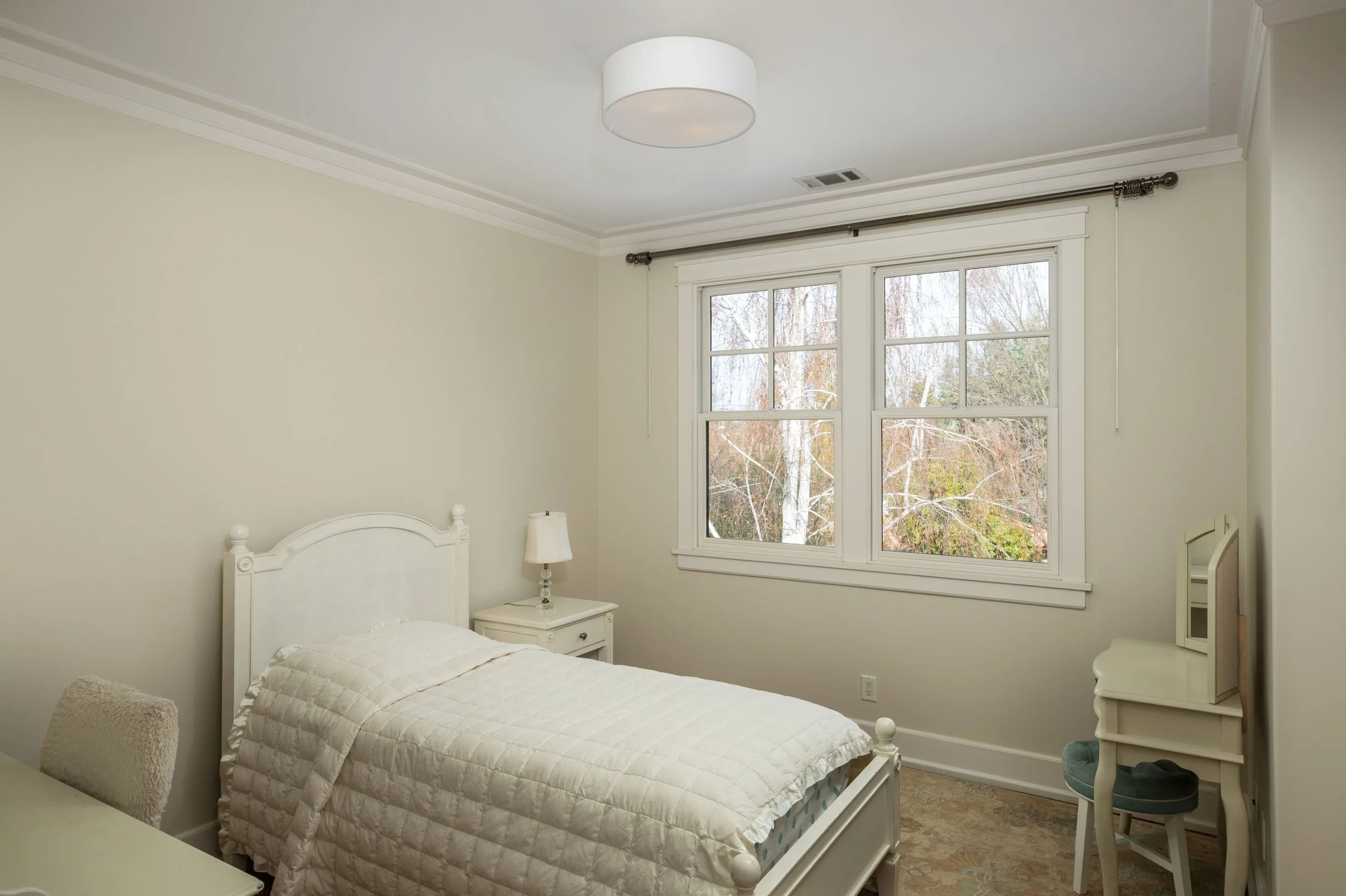 A bright bedroom with a white bed, nightstand, lamp, dresser, and a window showing trees outside.