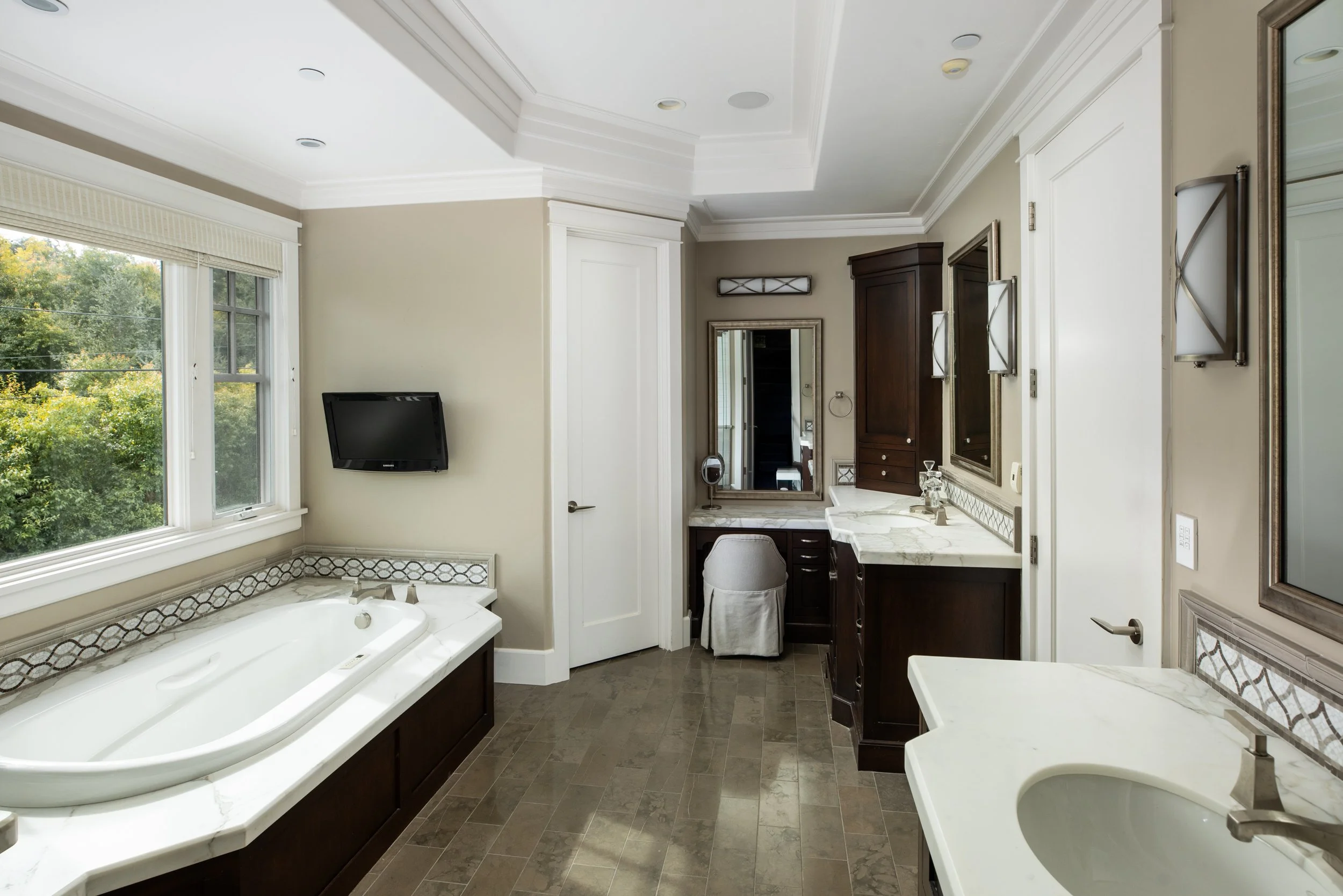 Bathroom with a bathtub, vanity with marble countertop, mirror, wall-mounted TV, and a window with a view of green trees.