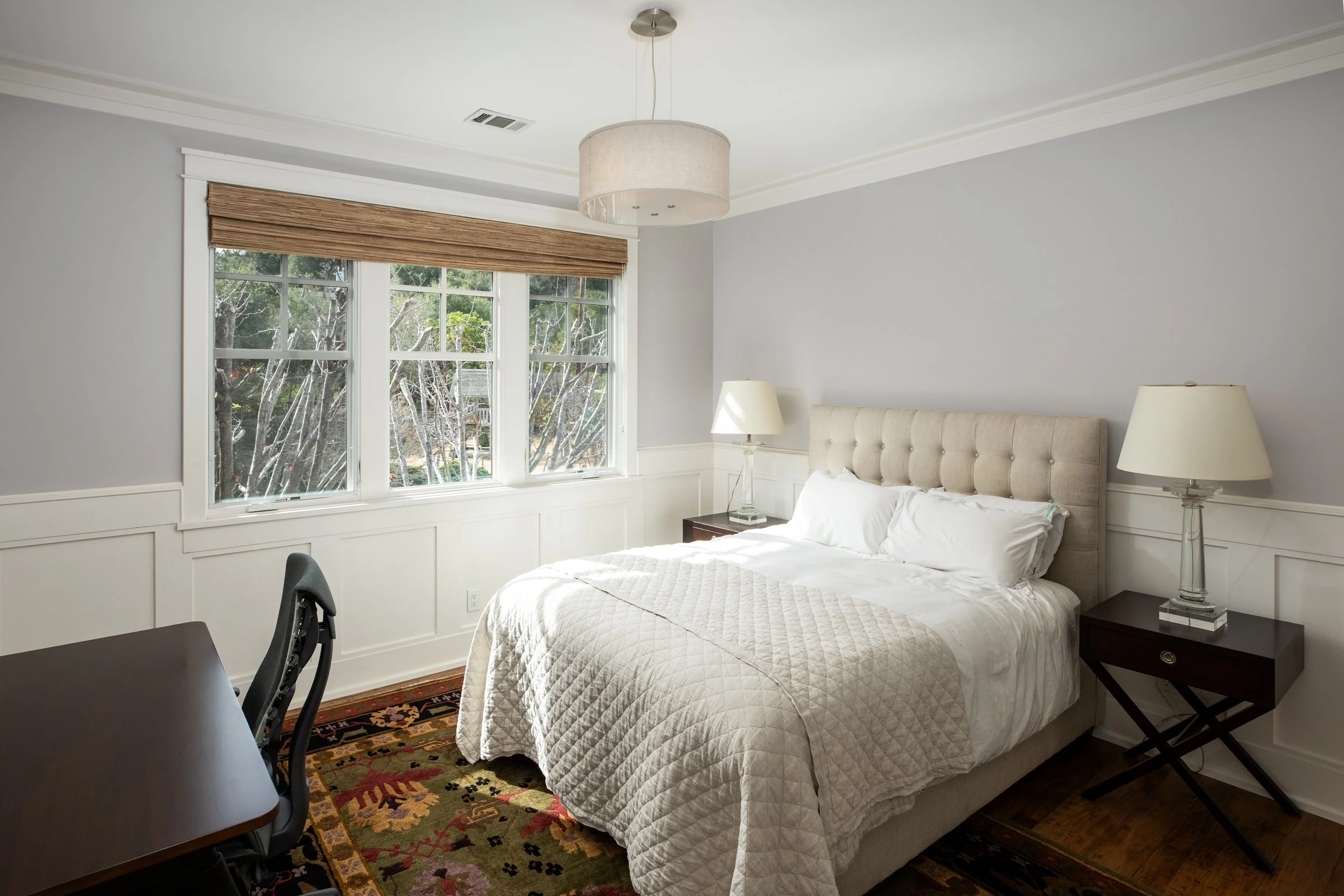 A bedroom with a king-size bed with white bedding and beige tufted headboard, two nightstands with lamps, and a window with brown blinds overlooking trees.