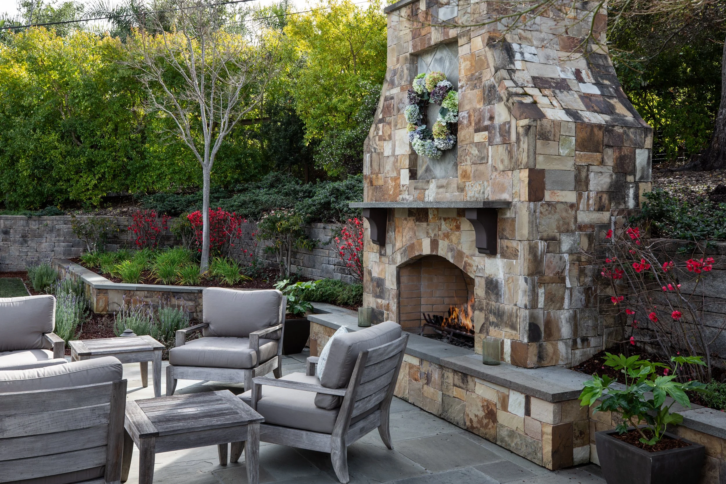 Outdoor patio with stone fireplace, wooden chairs with cushions, and surrounding landscaped garden with trees and flowering bushes.