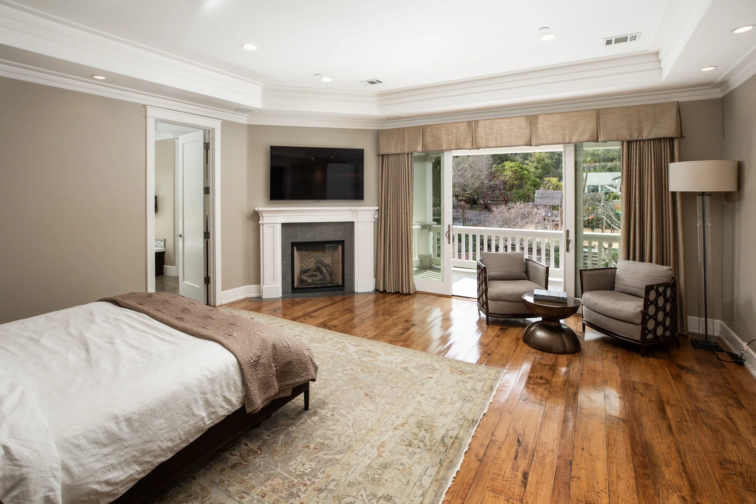 A bedroom with a bed, a fireplace with a mounted TV above, a sliding door leading to a balcony, two chairs and a small table, a floor lamp, and hardwood flooring.