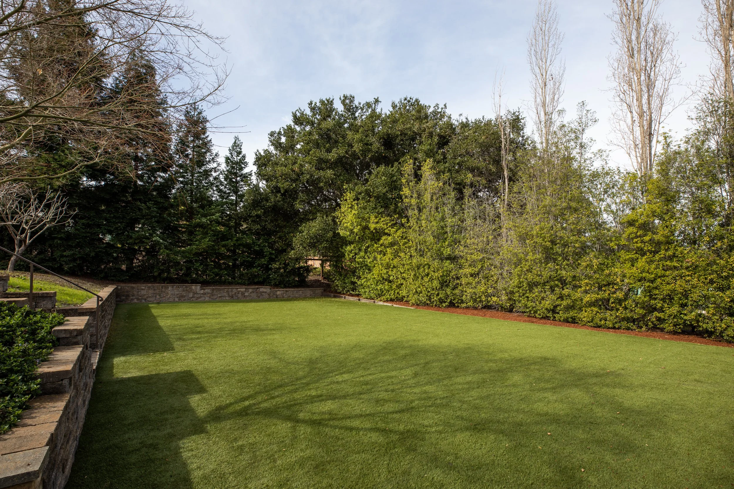 A well-maintained backyard with a lush green lawn, a stone retaining wall with steps on the left, and a variety of trees and bushes along the edges. The sky is partly cloudy.