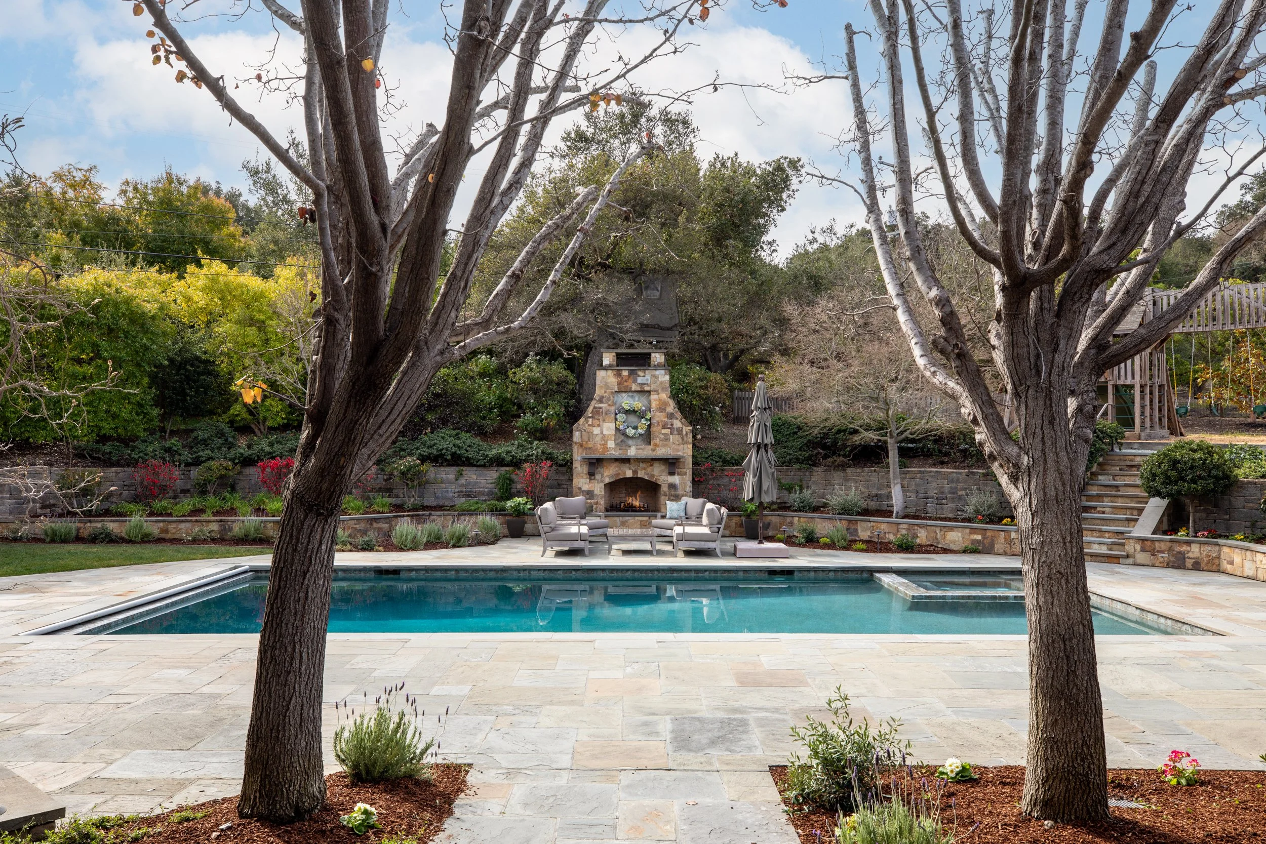A backyard with a swimming pool, outdoor seating area, stone fireplace, and trees in a landscaped yard.