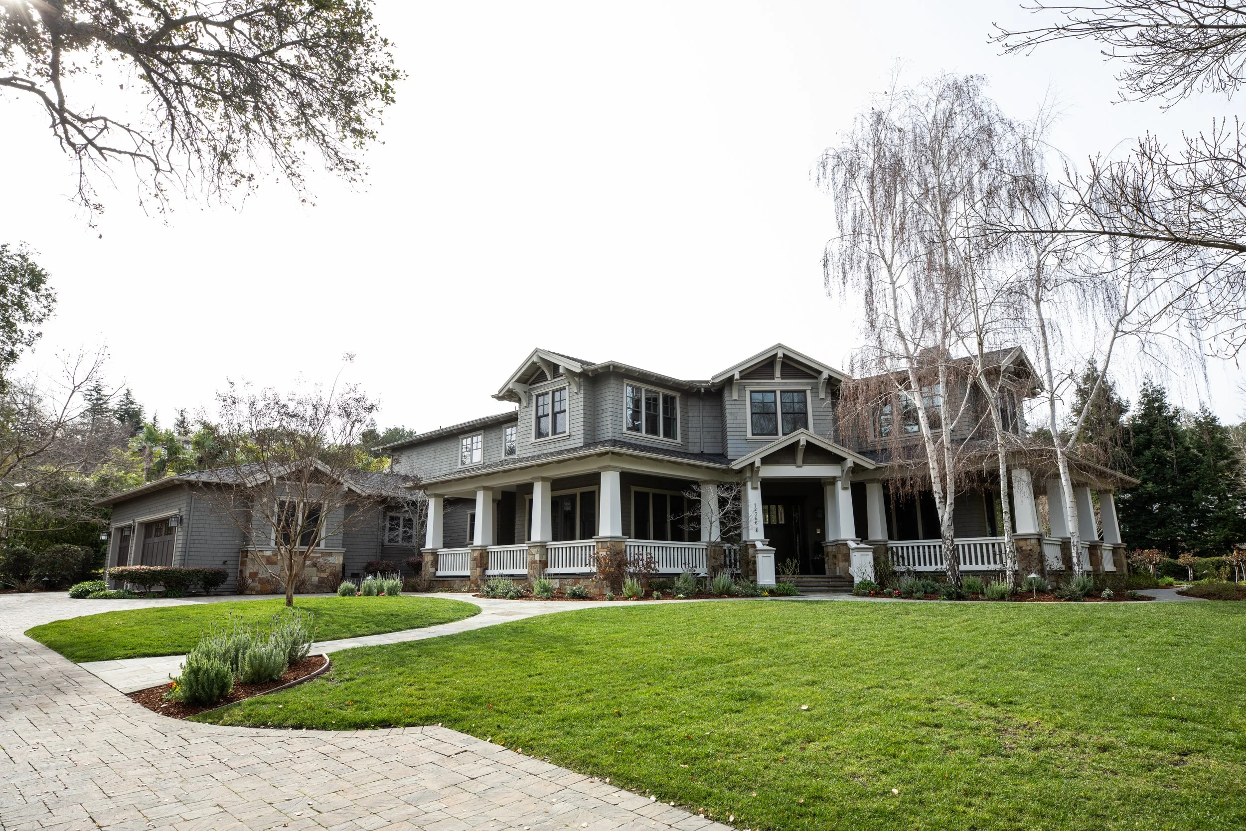 Large gray two-story house with a front porch, surrounded by a well-maintained lawn and trees, under an overcast sky.