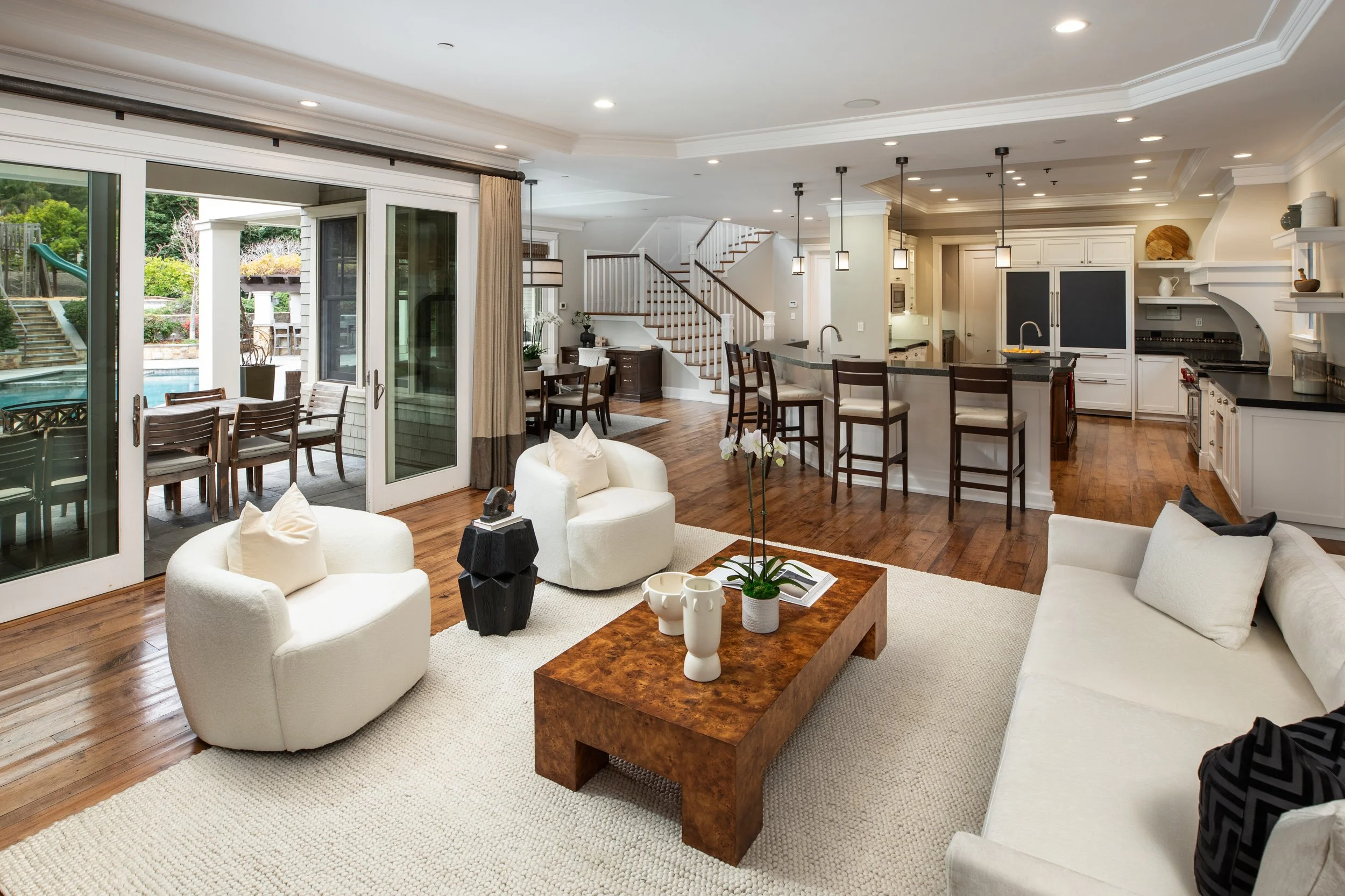 Spacious open-concept living room and kitchen with hardwood floors, white furniture, a wooden coffee table, and large sliding glass doors leading to an outdoor patio with a pool and stairs.