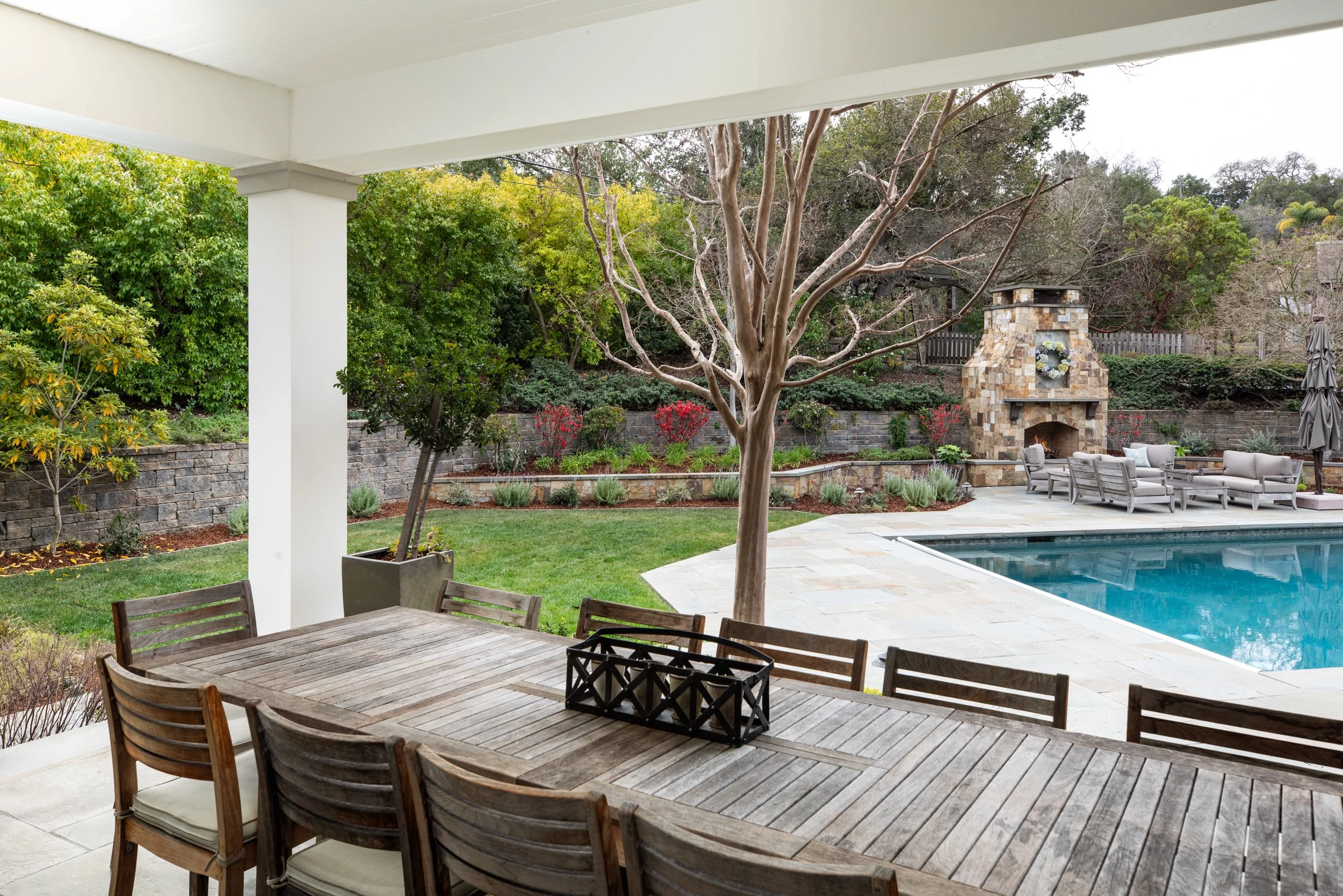 View of backyard patio area with dining table, tree, grass, lush green bushes, stone retaining wall, outdoor fireplace, pool with lounge chairs, and covered patio