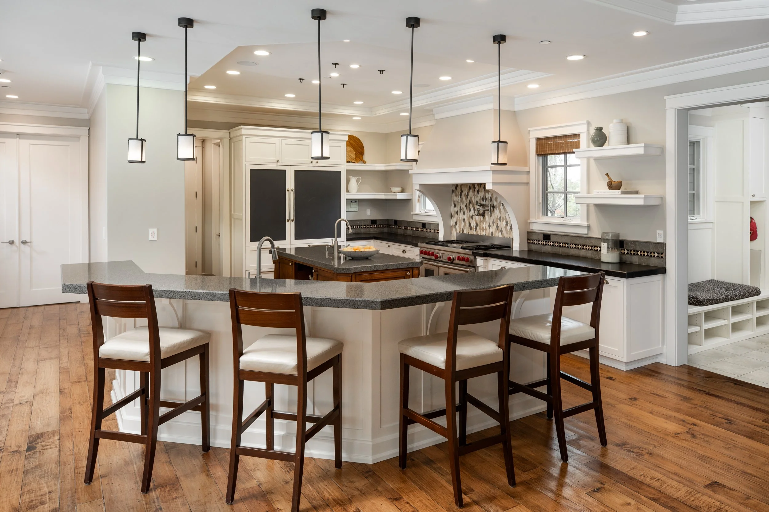 Modern kitchen with white cabinets, black countertops, a central island, and wooden flooring go to the ceiling with hanging pendant lights. There are bar stools around the island, a window above the sink, and decorative items on open shelves.