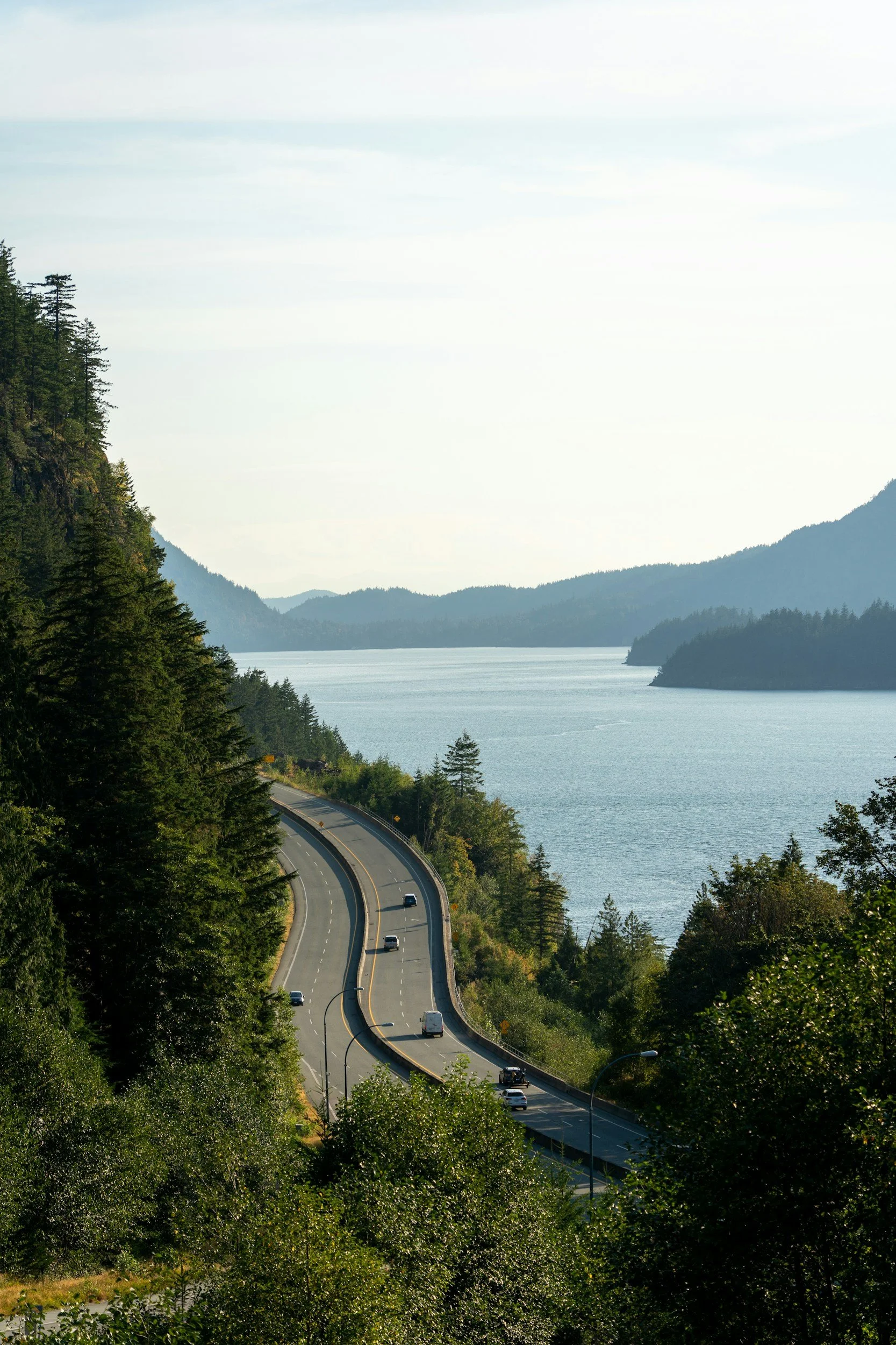 A winding coastal road runs between dense green trees and a large body of water, with mountains in the background under a partly cloudy sky.