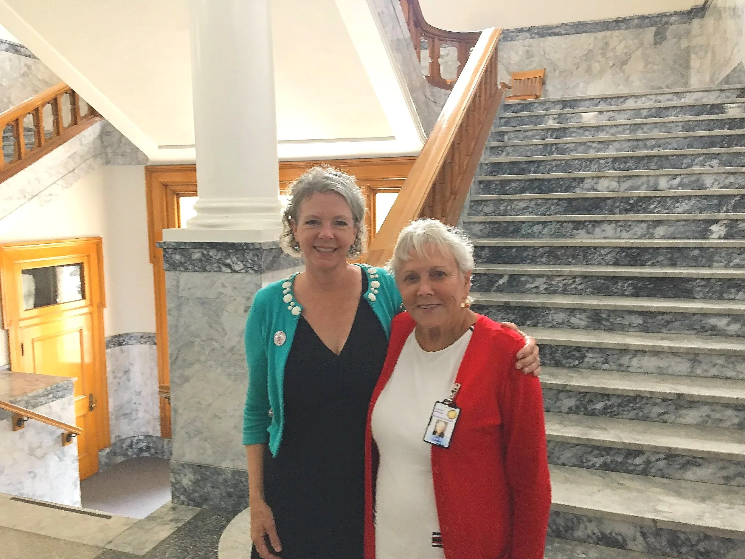 Lisa and Friend smiling and standing close together in front of a marble staircase inside wasco county courthouse in The Dalles with wood trim and large windows.