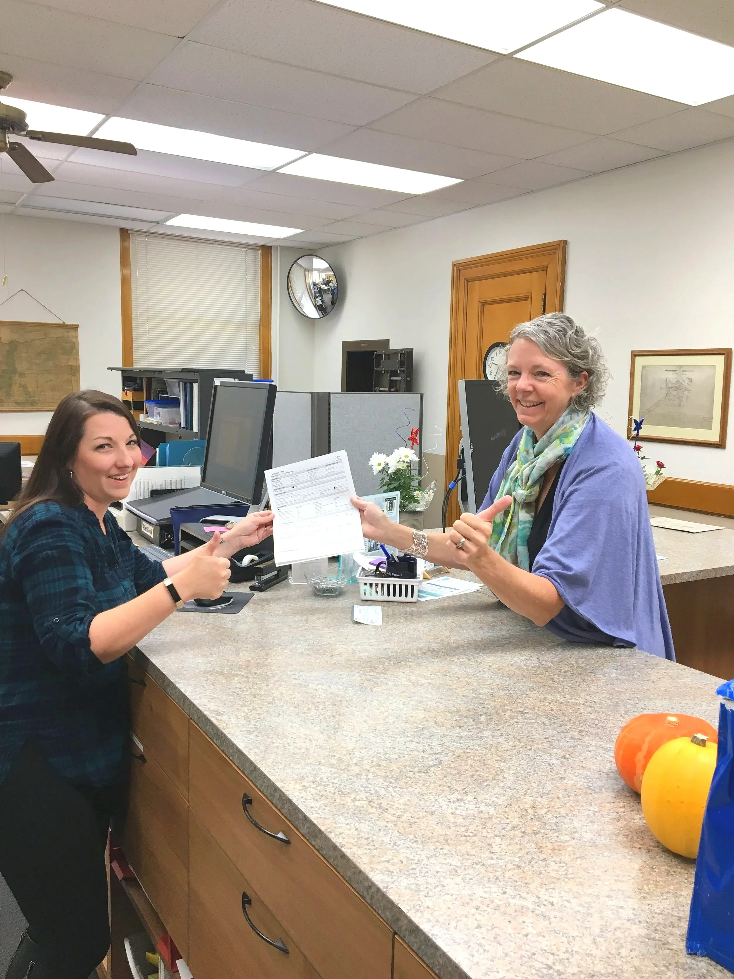 Lisa Gambee At Wasco County Filing . One woman, with dark hair, is handing a document to the other woman, who has gray hair and is smiling and pointing at the document. The office has computers, a clock, plants, and framed pictures on the walls.