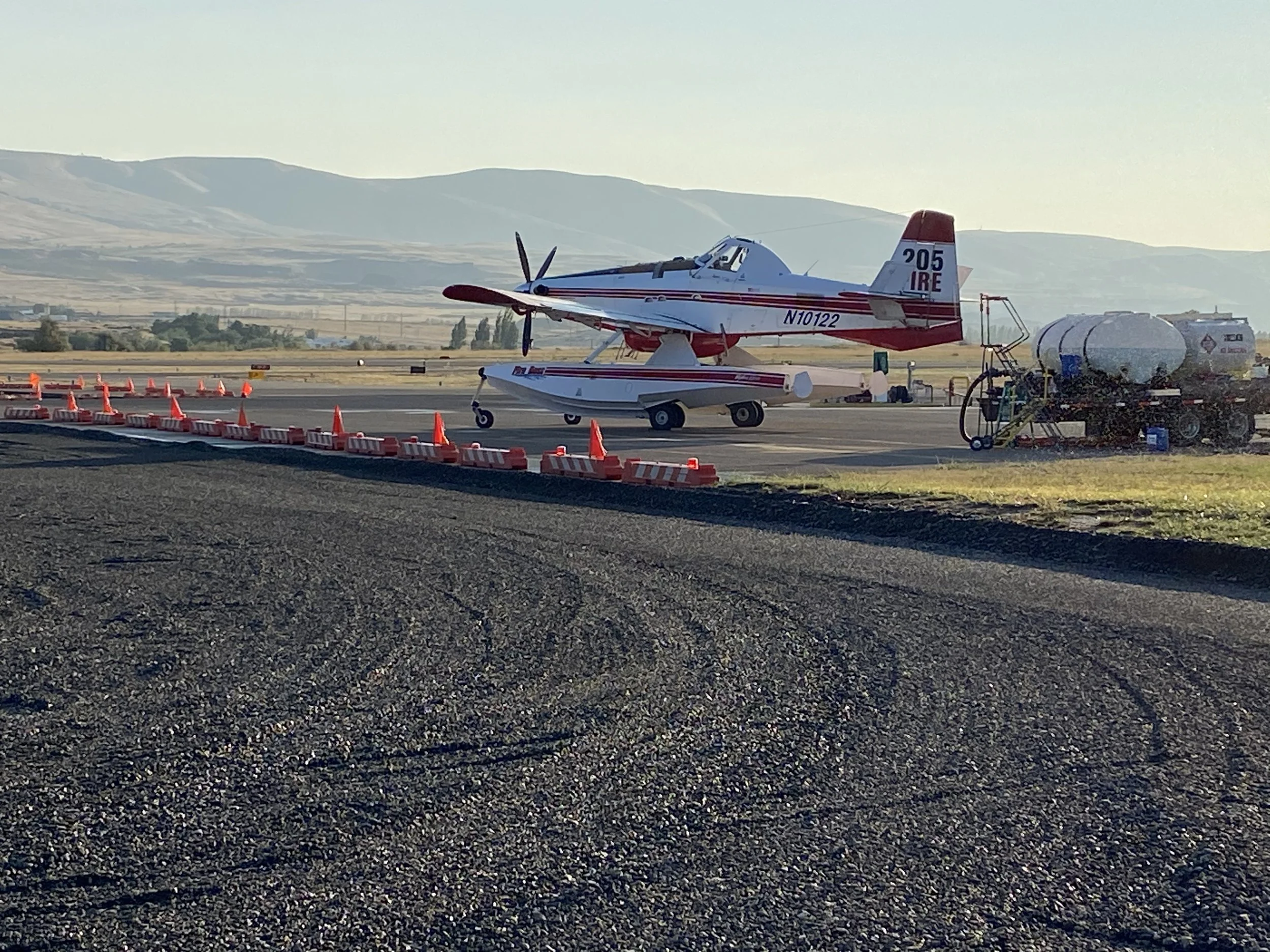 Fire scooper plane with a white and red body parked on an airfield, with a water tanker truck nearby and orange traffic cones and barriers on the ground.
