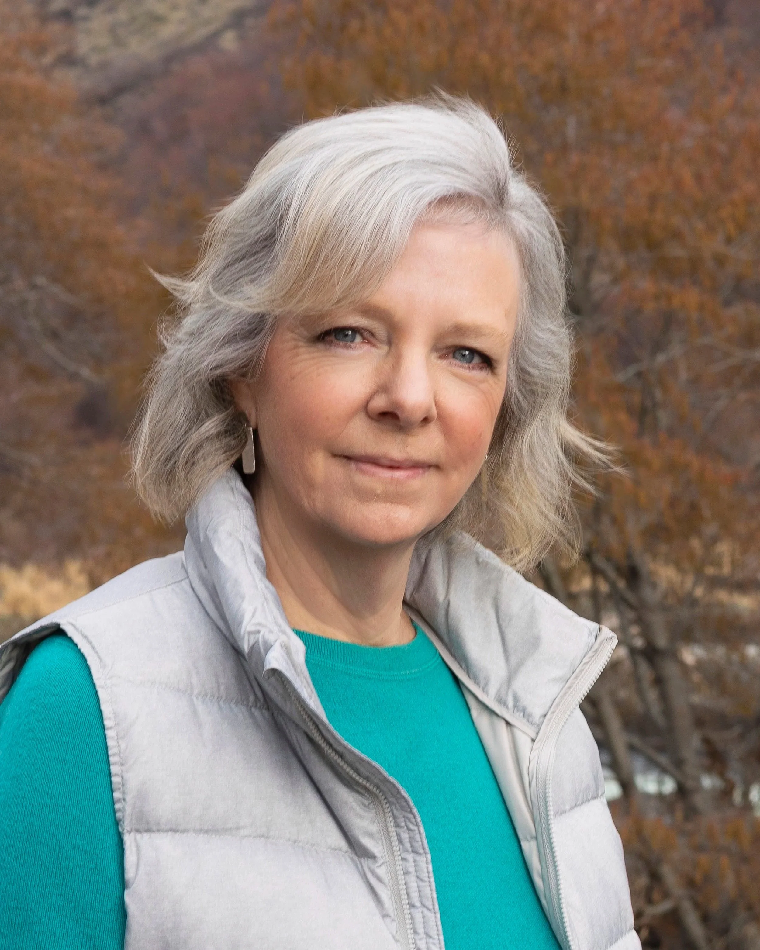 A woman with shoulder-length gray hair wearing a turquoise shirt and a light gray vest, standing outdoors with autumn-colored trees in the background.